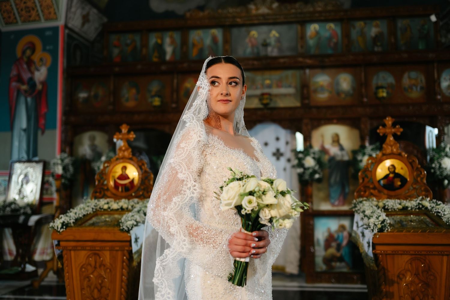 Bride in lace wedding dress and veil holding white bouquet inside ornate Orthodox church with icons.