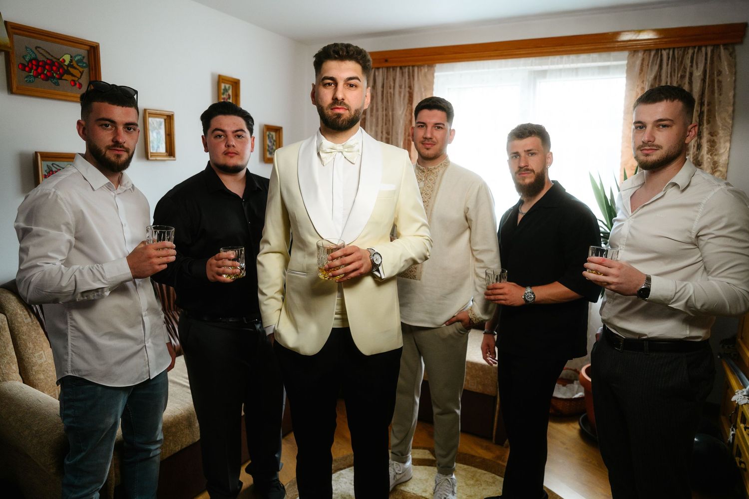 Groom in white tuxedo poses with groomsmen holding drinks in a decorated indoor setting.