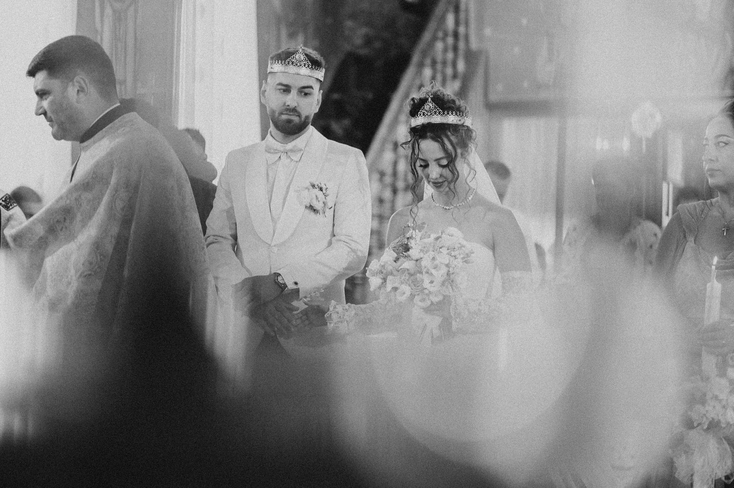 Black and white wedding ceremony photo of bride and groom wearing crowns standing before a priest in ornate church.