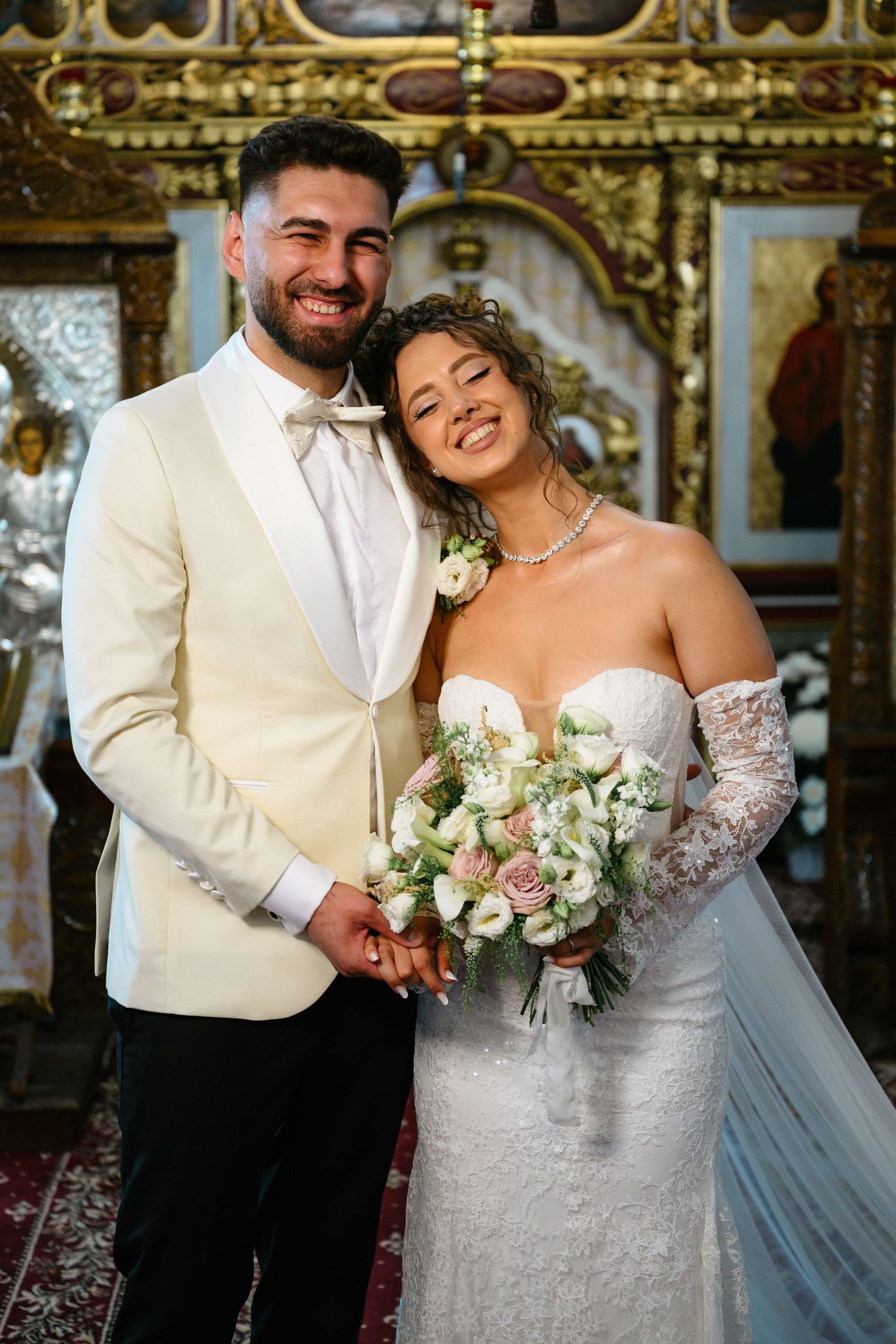 Smiling newlyweds pose in ornate church, bride in lace gown holding pink bouquet, groom in cream tuxedo.