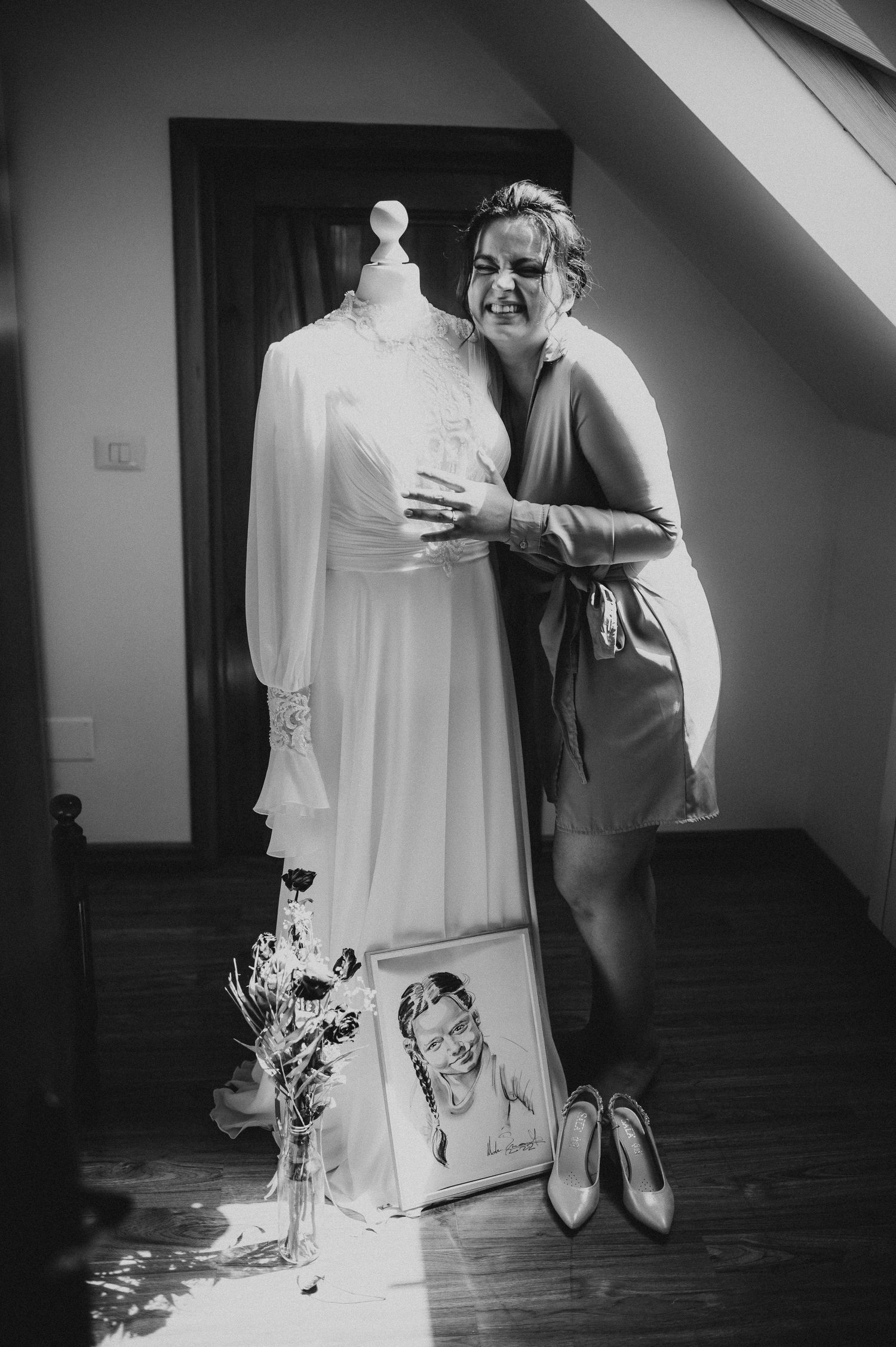 Bride in robe embracing white long-sleeve wedding gown on mannequin in black and white photo.