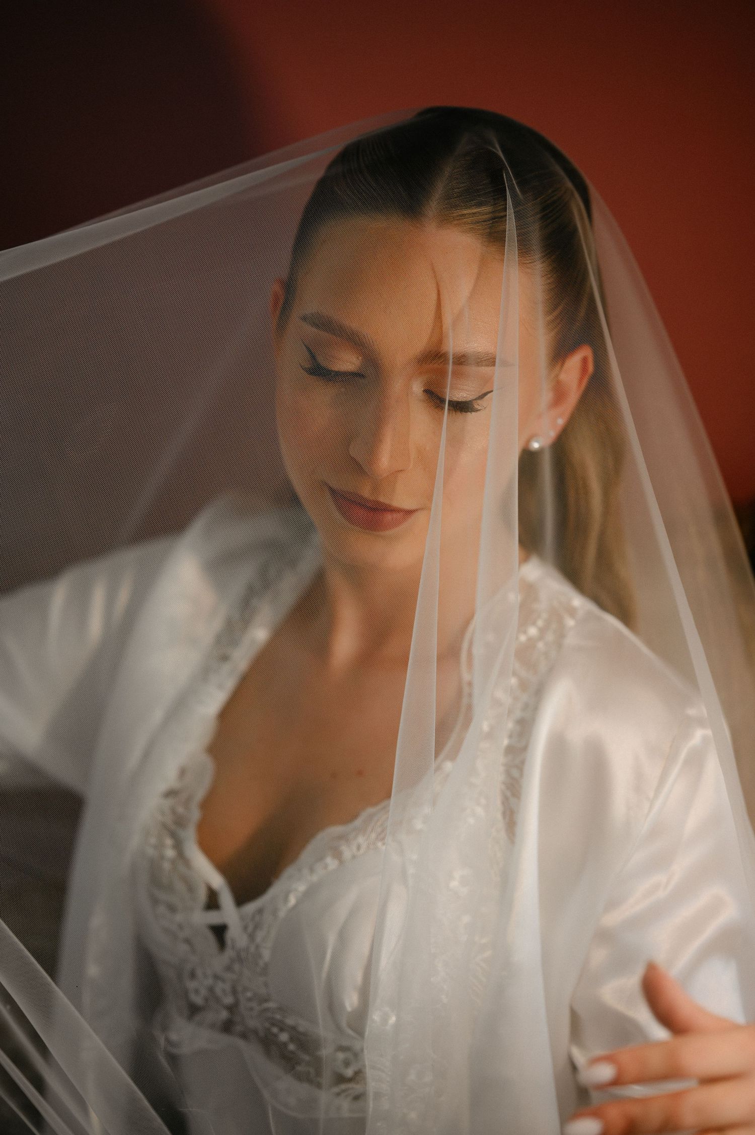 Bride in white satin robe with veil draped over head, eyes closed.