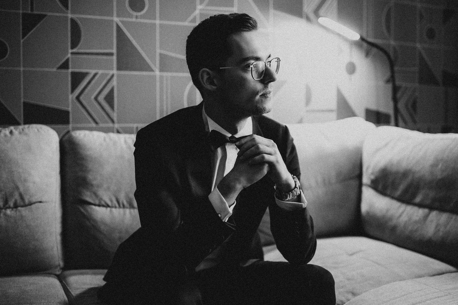 Black and white photo of a stylish man in a suit and sunglasses sitting on a couch in a modern room.