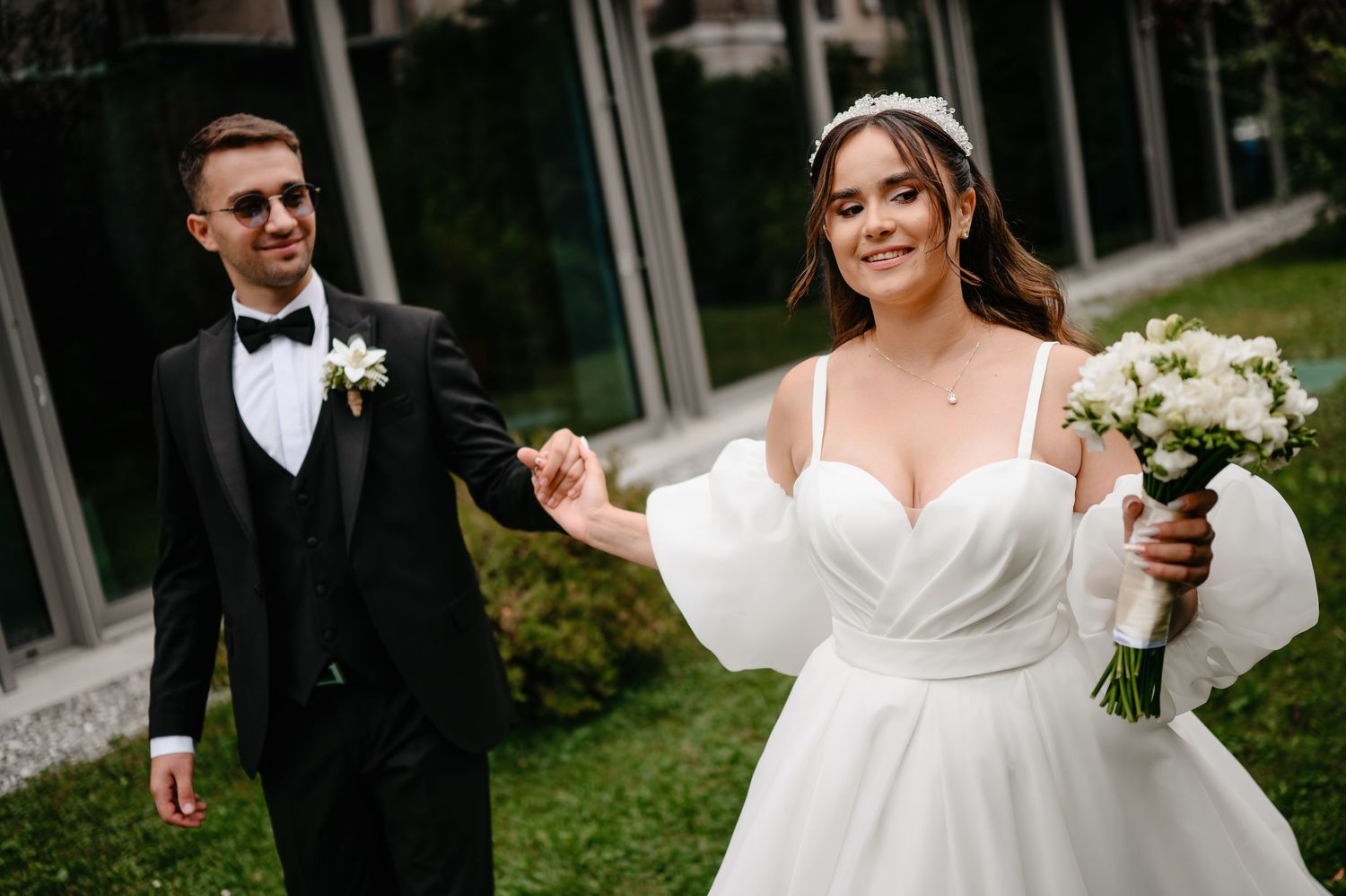 Smiling bride in white off-shoulder gown holding bouquet walks with groom in black tuxedo outdoors.