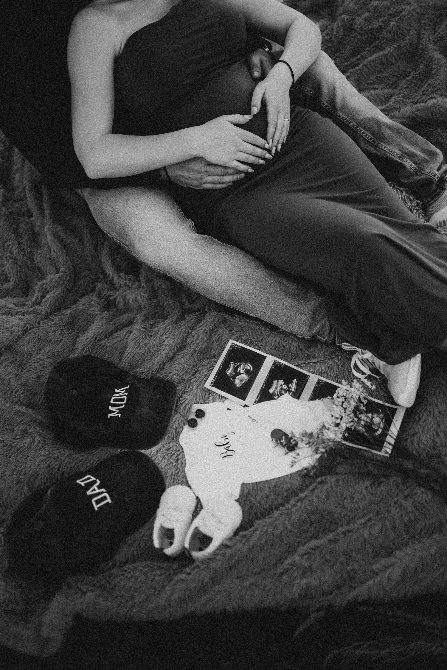 Black and white overhead shot of pregnant couple with ultrasound photos and baby items on bed.