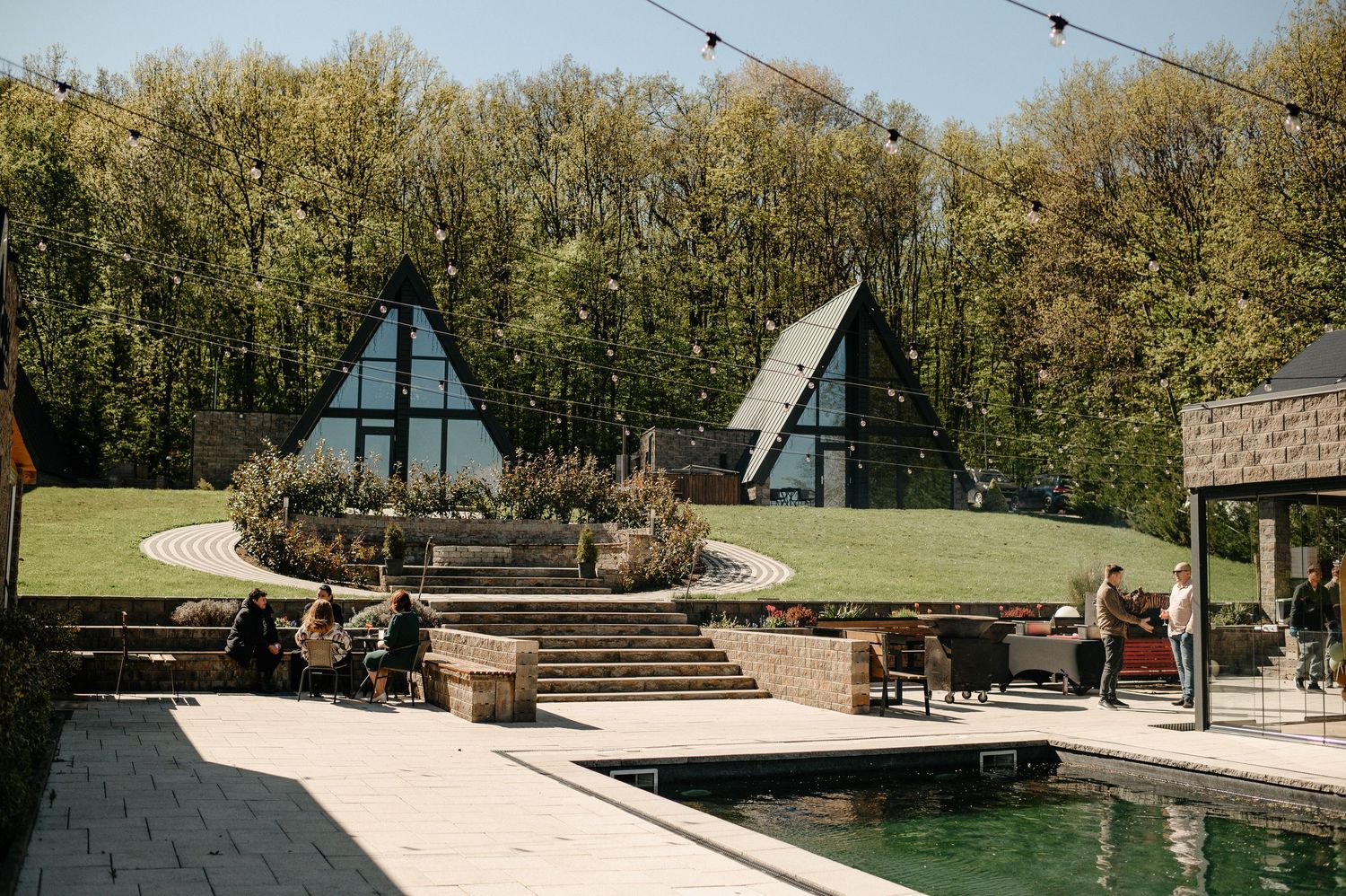 Modern A-frame cabin retreat with pond, terrace seating, and lush green landscape.