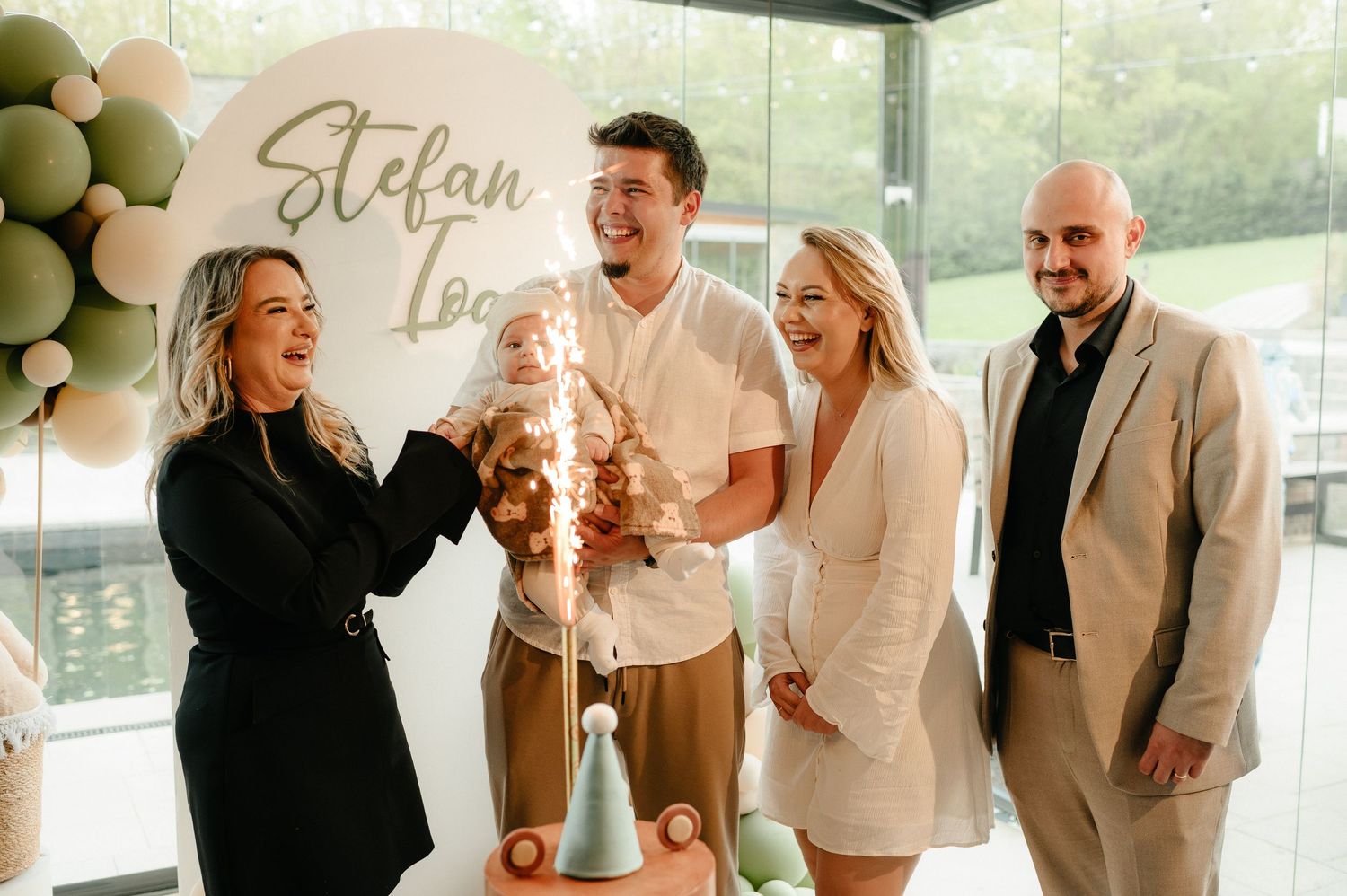 Joyful family group celebrating baby Stefan Ioan's christening with sparkler cake and balloon backdrop.