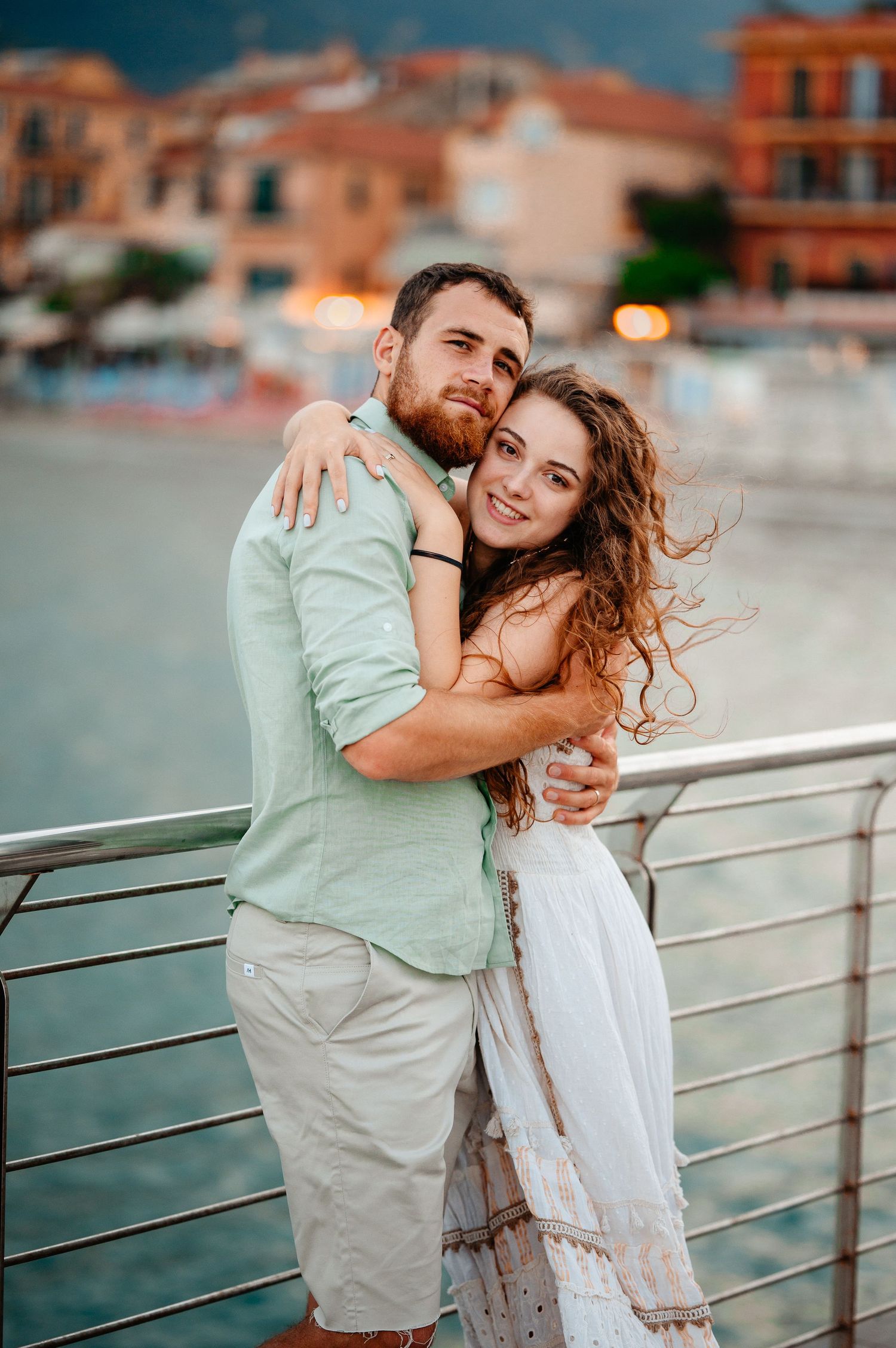 Loving couple embracing on waterfront promenade with colorful coastal town behind them.