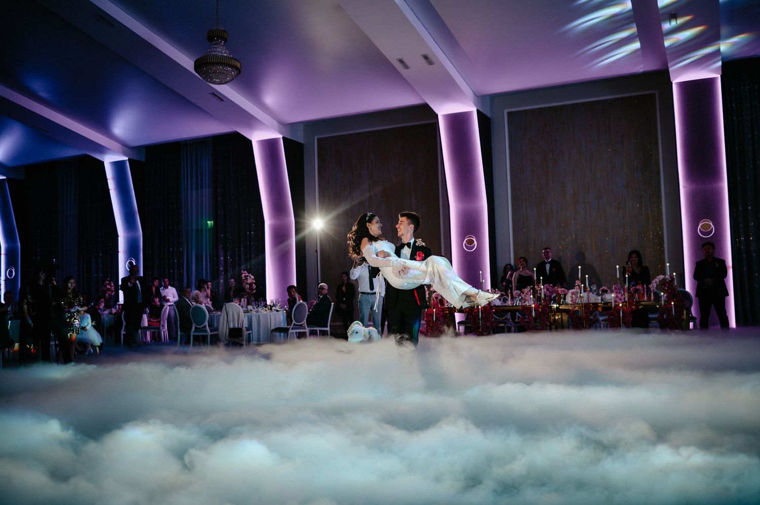 Groom carries bride in white dress during magical first dance enveloped in cloud-like dry ice fog.