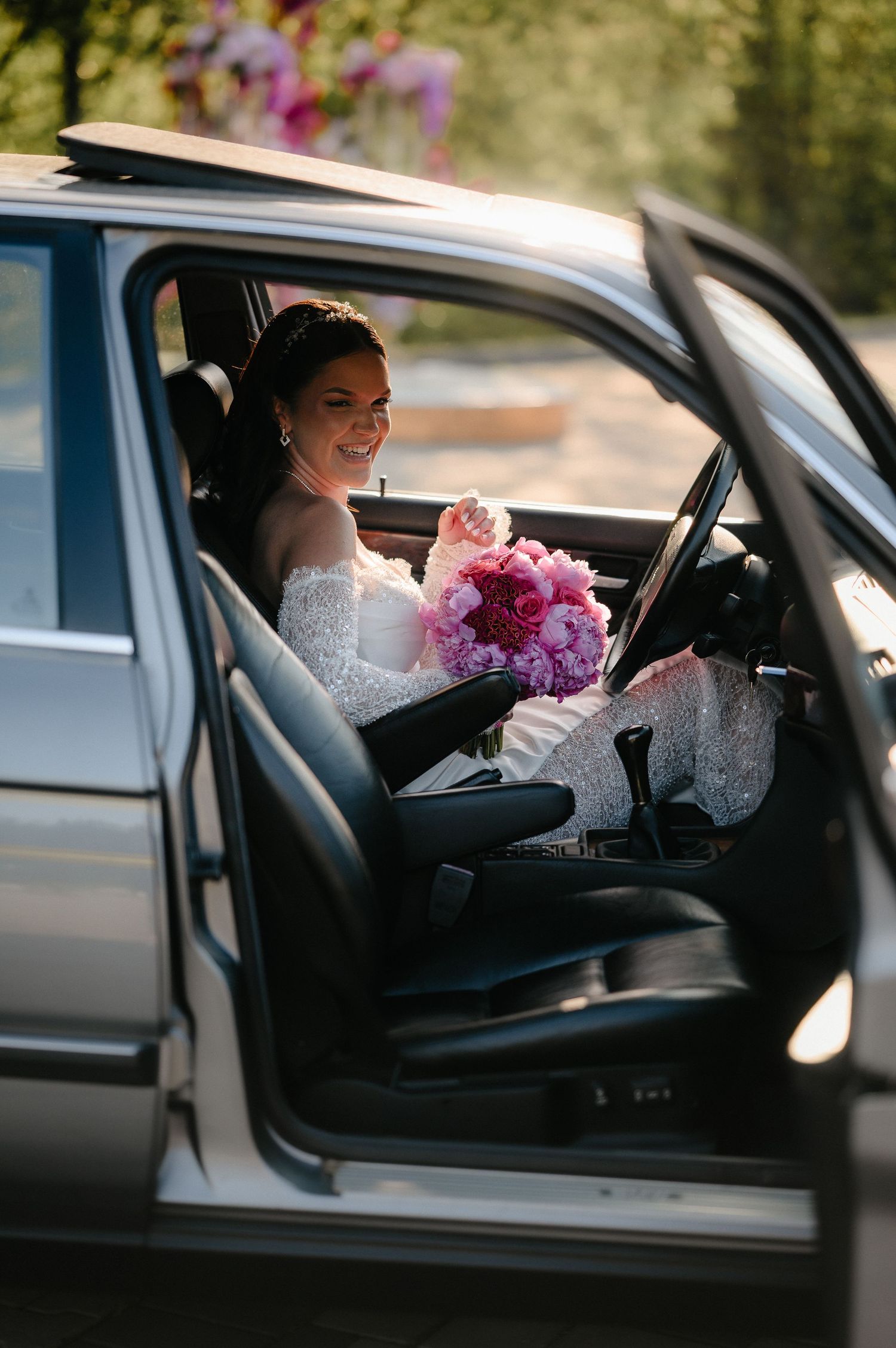 Radiant bride in off-shoulder lace gown smiles while seated in vintage car with pink floral bouquet.