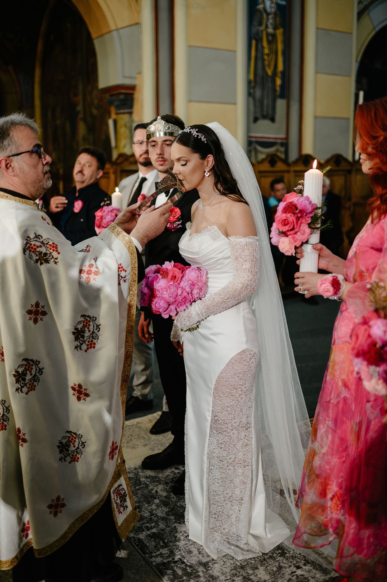 Bride in white gown receives blessing from Orthodox priest holding candle during church ceremony.