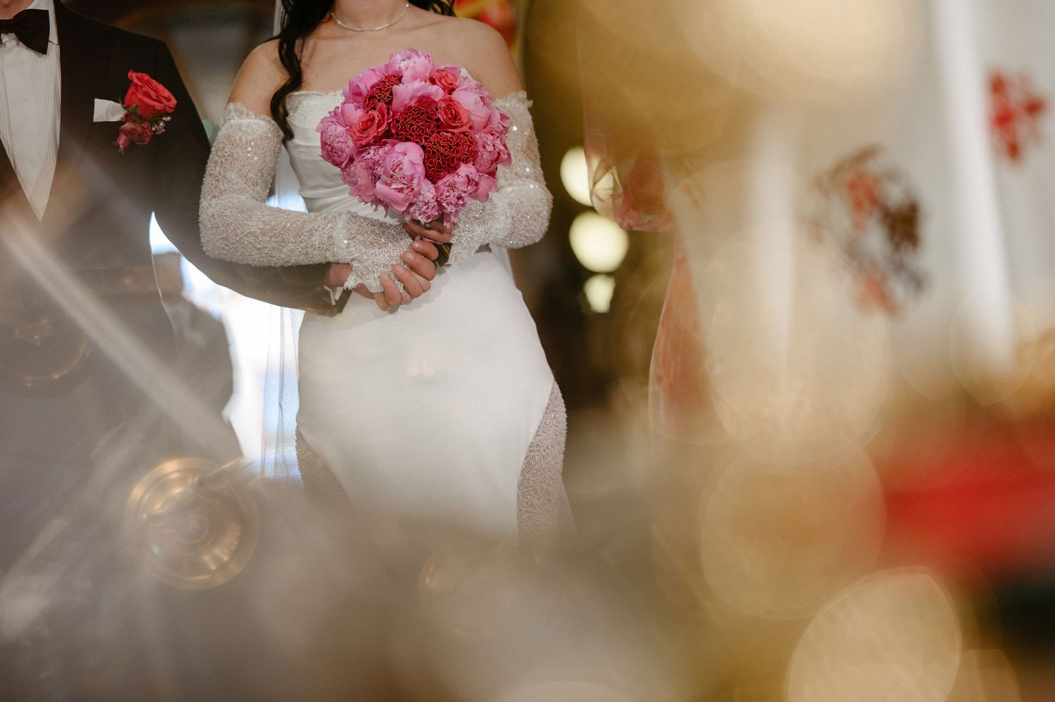 Close-up of bride in lace wedding gown holding vibrant pink peony bouquet during church ceremony.