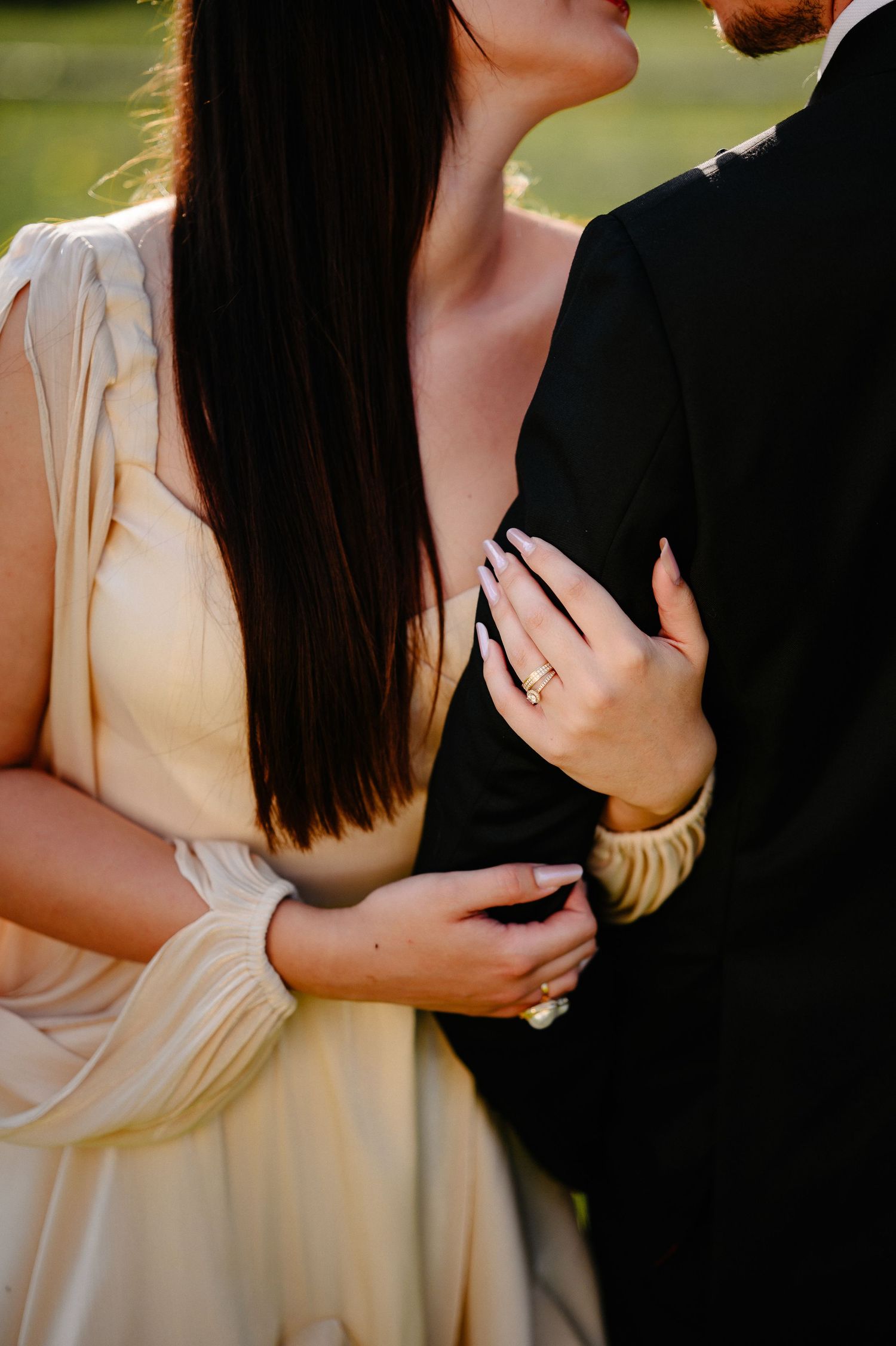 Close-up of a bride in cream dress resting her hand on a grooms black suit, showing engagement ring.