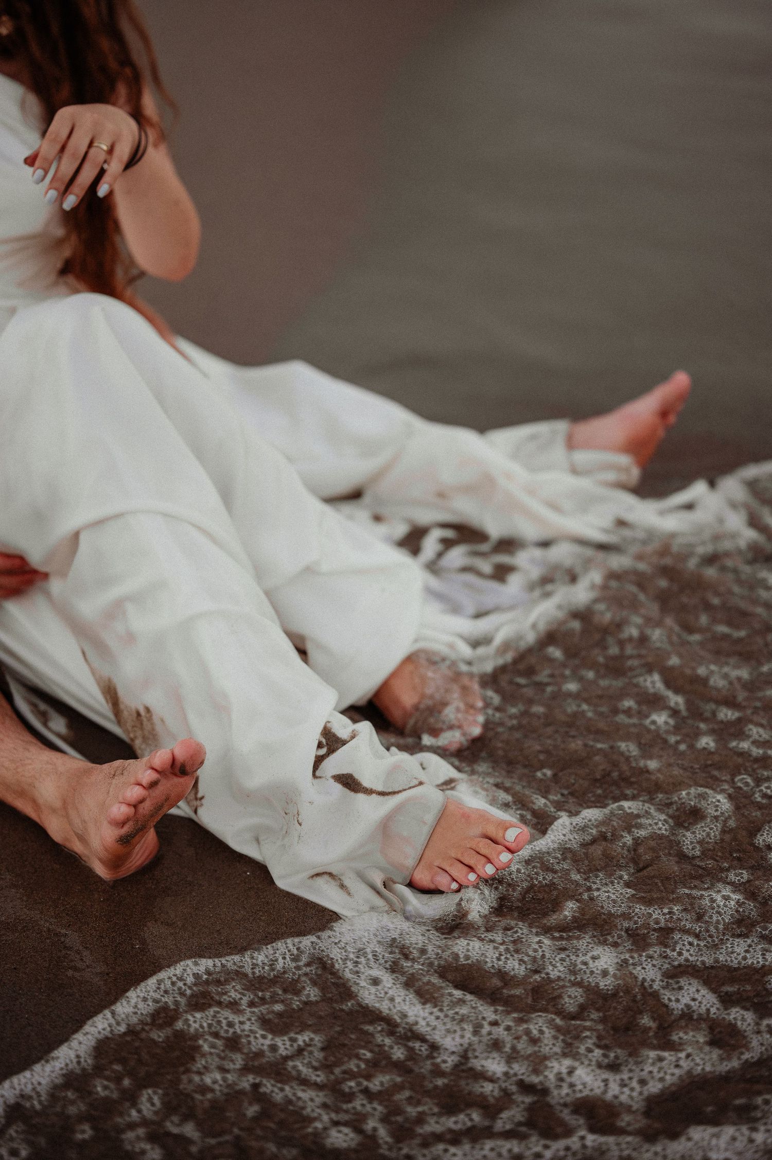 Bride in white wedding dress sitting barefoot on wet sand with waves washing over feet at beach.
