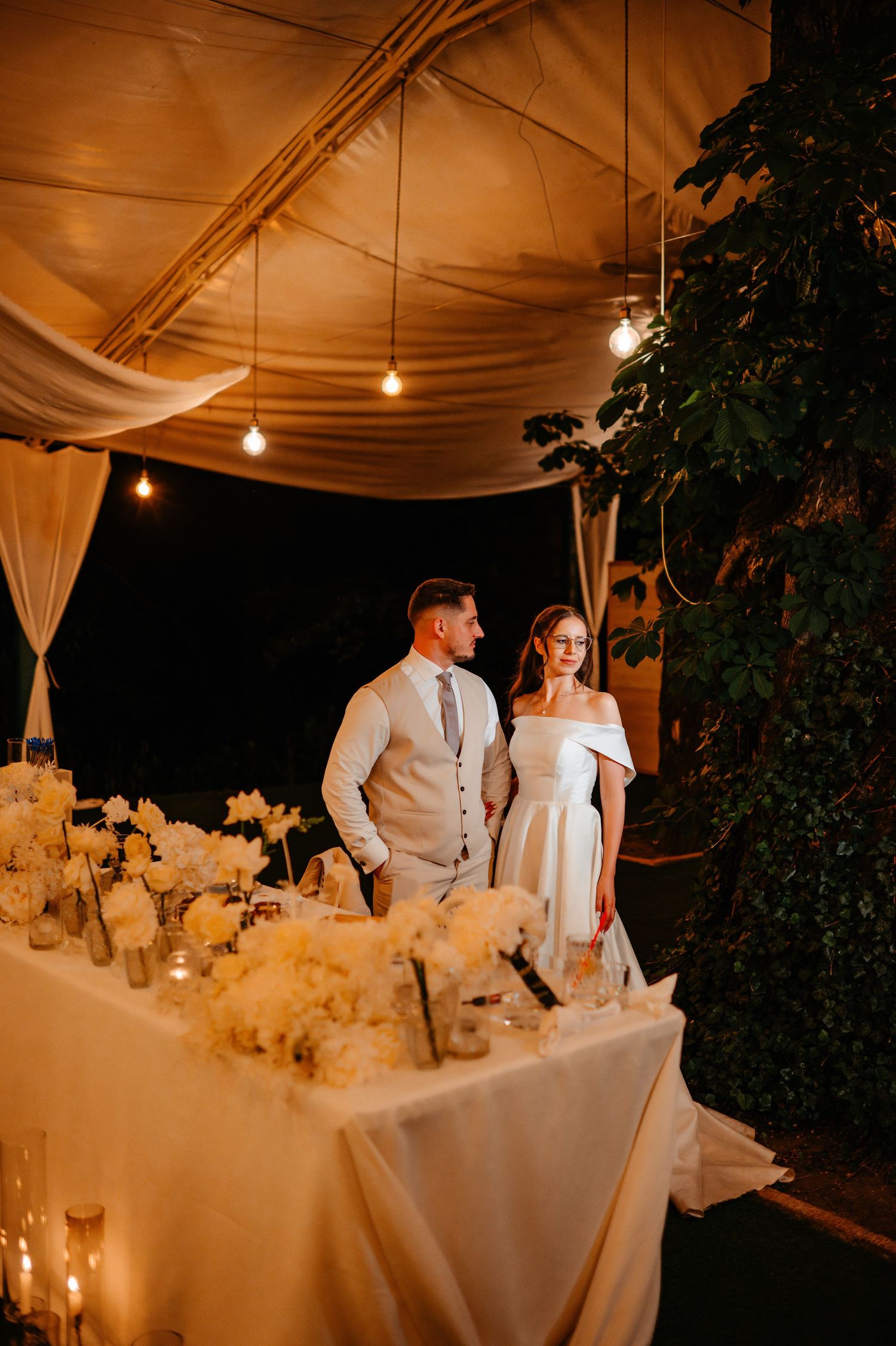 Bride and groom standing at elegantly decorated sweetheart table with white florals under draped tent at night.