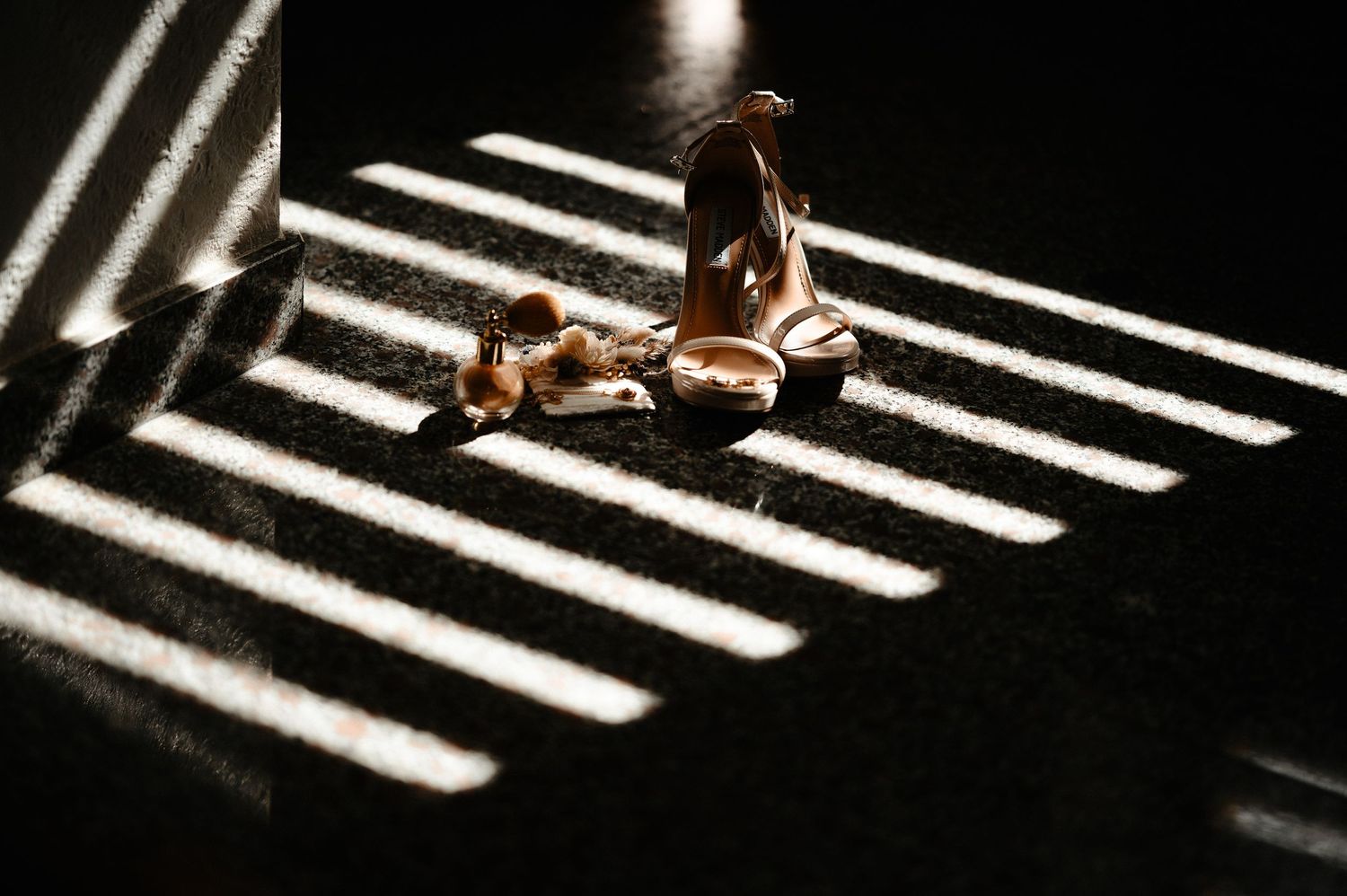 Gold bridal heels cast dramatic striped shadows on dark floor in artistic wedding photography.