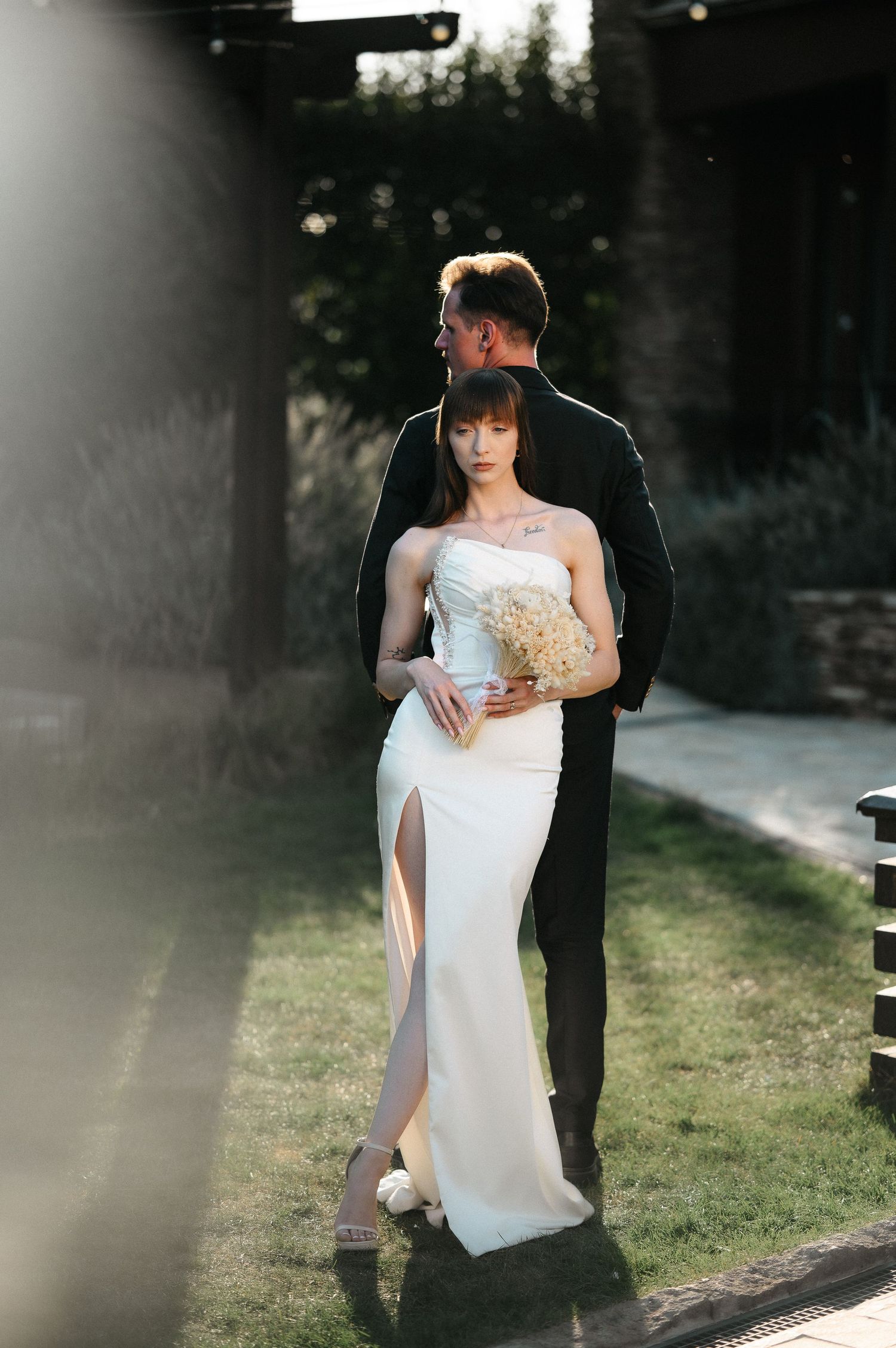Bride in white gown holding bouquet embraced by groom in black suit outdoors in golden sunlight.
