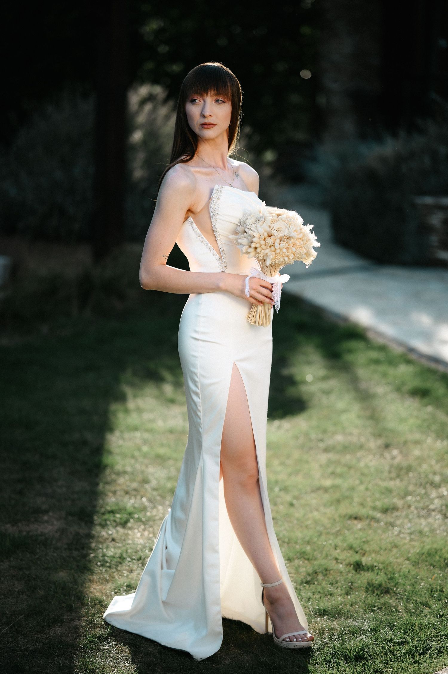 Elegant bride in a white slit gown holding a bouquet while posing in a sunlit garden.