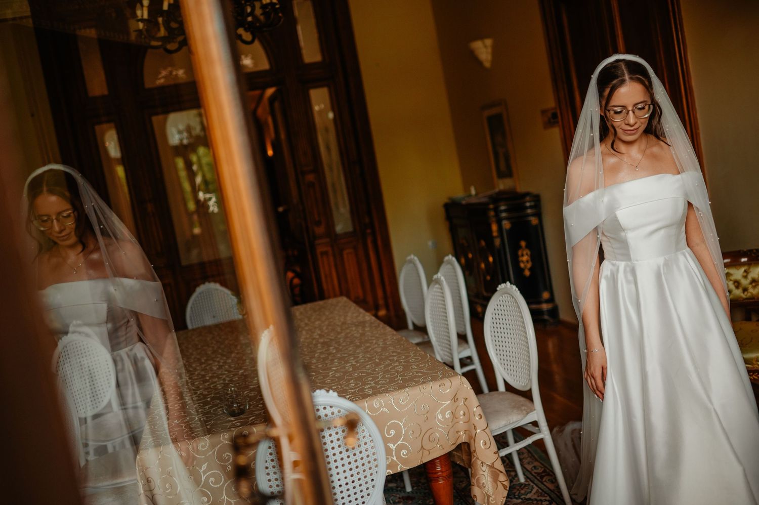Bride in white off-shoulder gown and veil reflected in mirror beside elegant dining room.