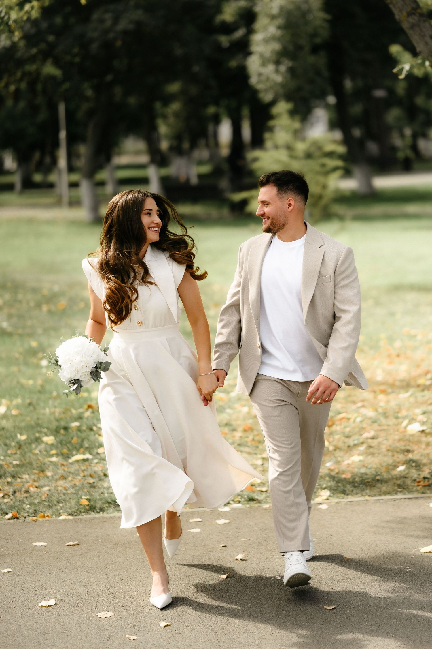 Happy couple walks hand-in-hand in sunny park, bride in white dress holding bouquet, groom in beige suit smiling.