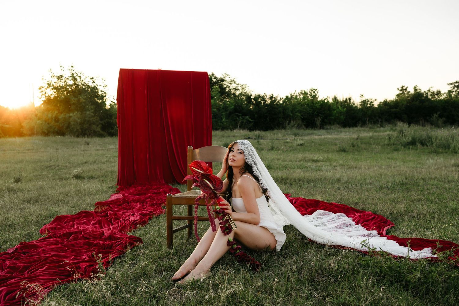 Bride in white lingerie and veil sits in sunlit field with red velvet backdrop and fabric at golden hour.