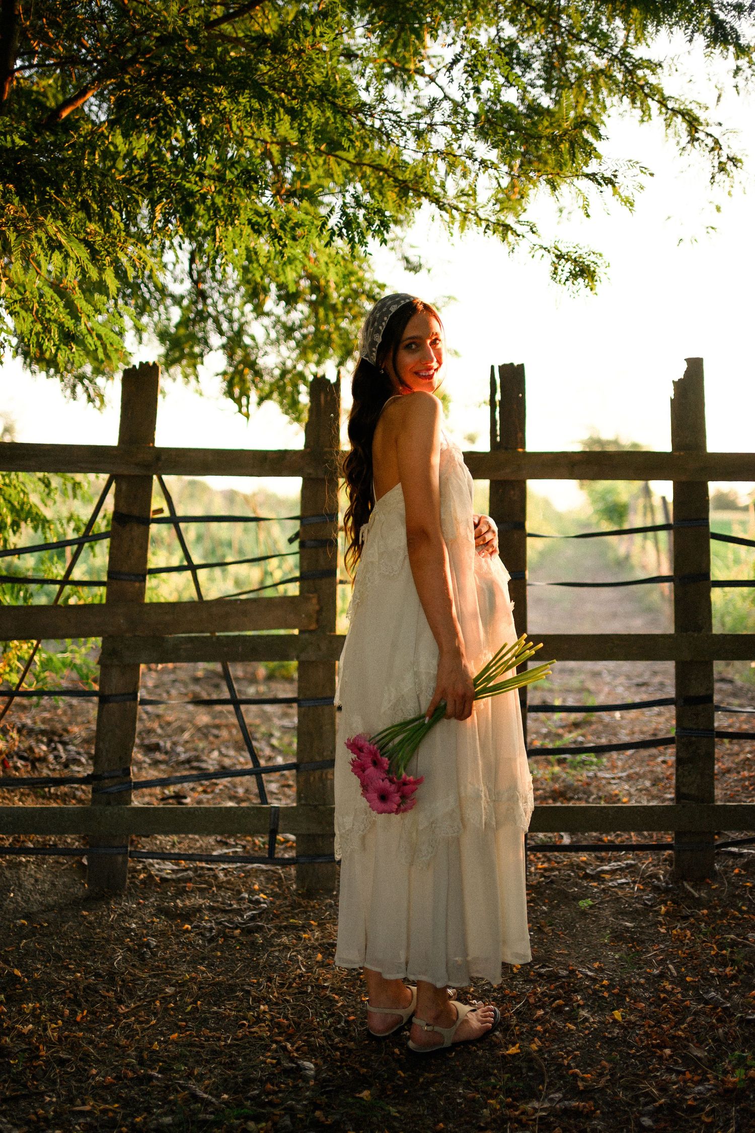 Young woman in white dress holding pink tulips smiles over shoulder by rustic wooden fence in golden sunlight.
