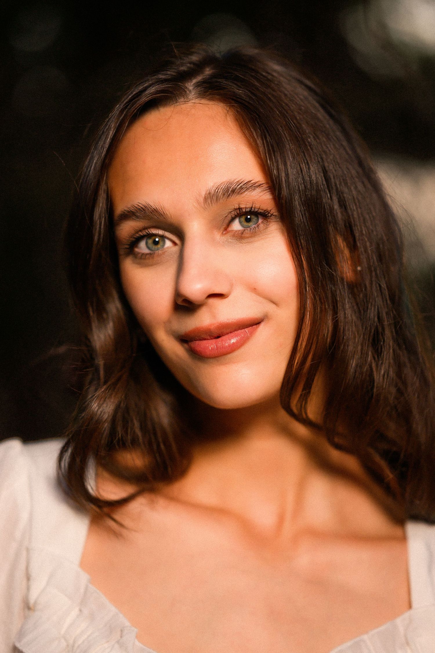 Young woman with dark wavy hair and green eyes smiling softly, wearing a white top in warm golden light.