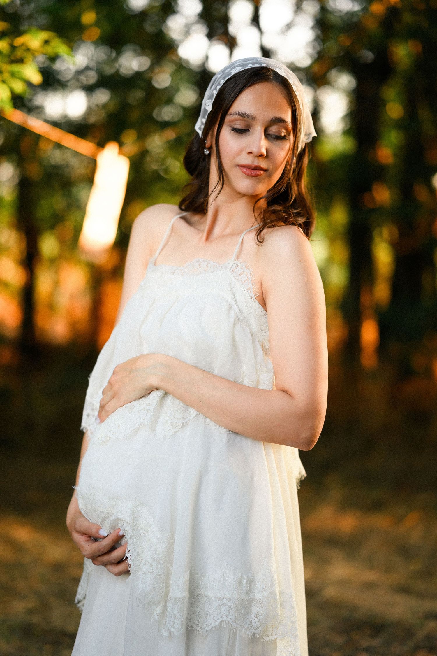 Pregnant woman in white lace dress and headpiece cradling her baby bump outdoors at golden hour.