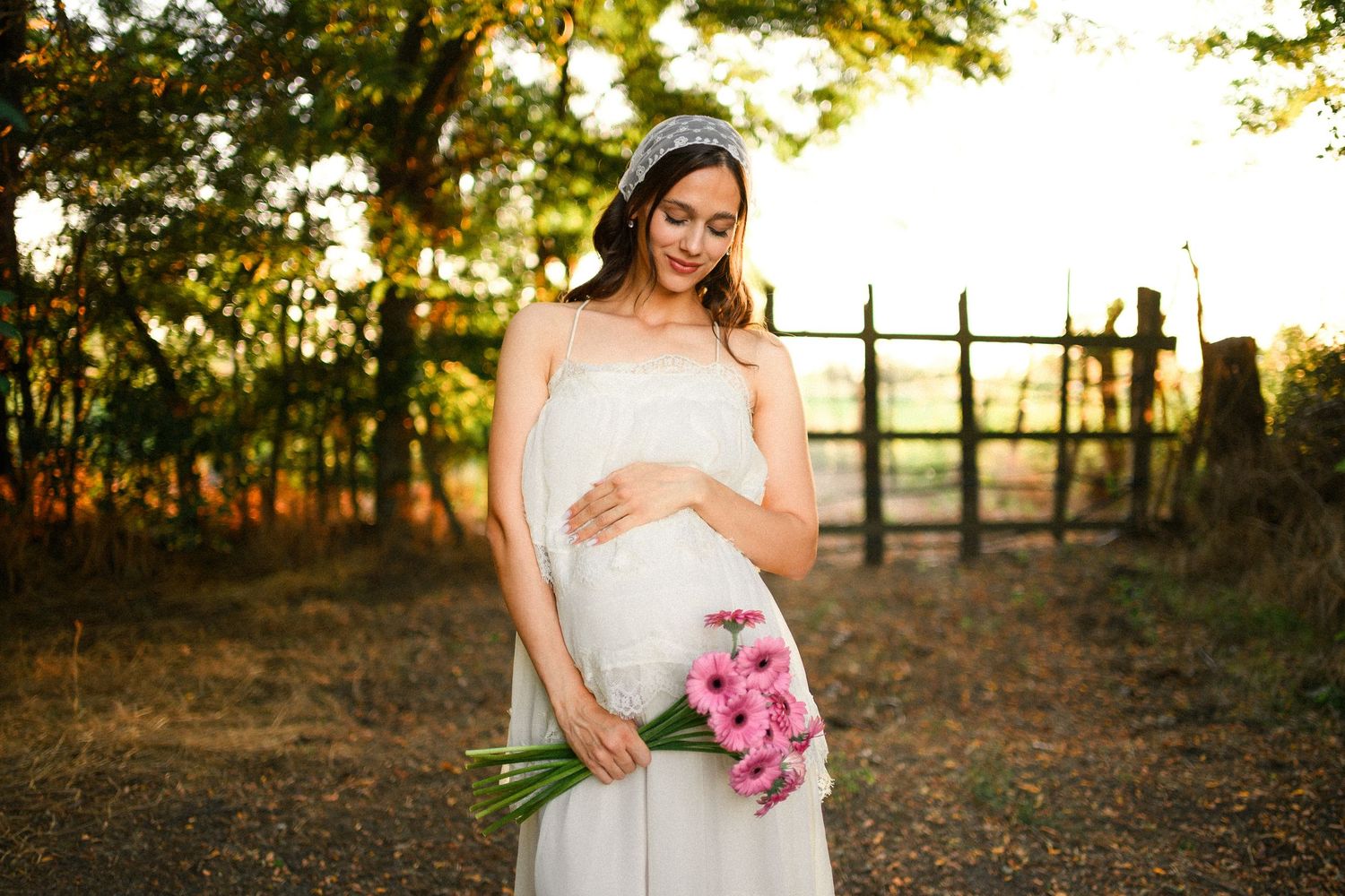 Pregnant woman in white dress holding pink gerbera bouquet outdoors in golden sunlight.