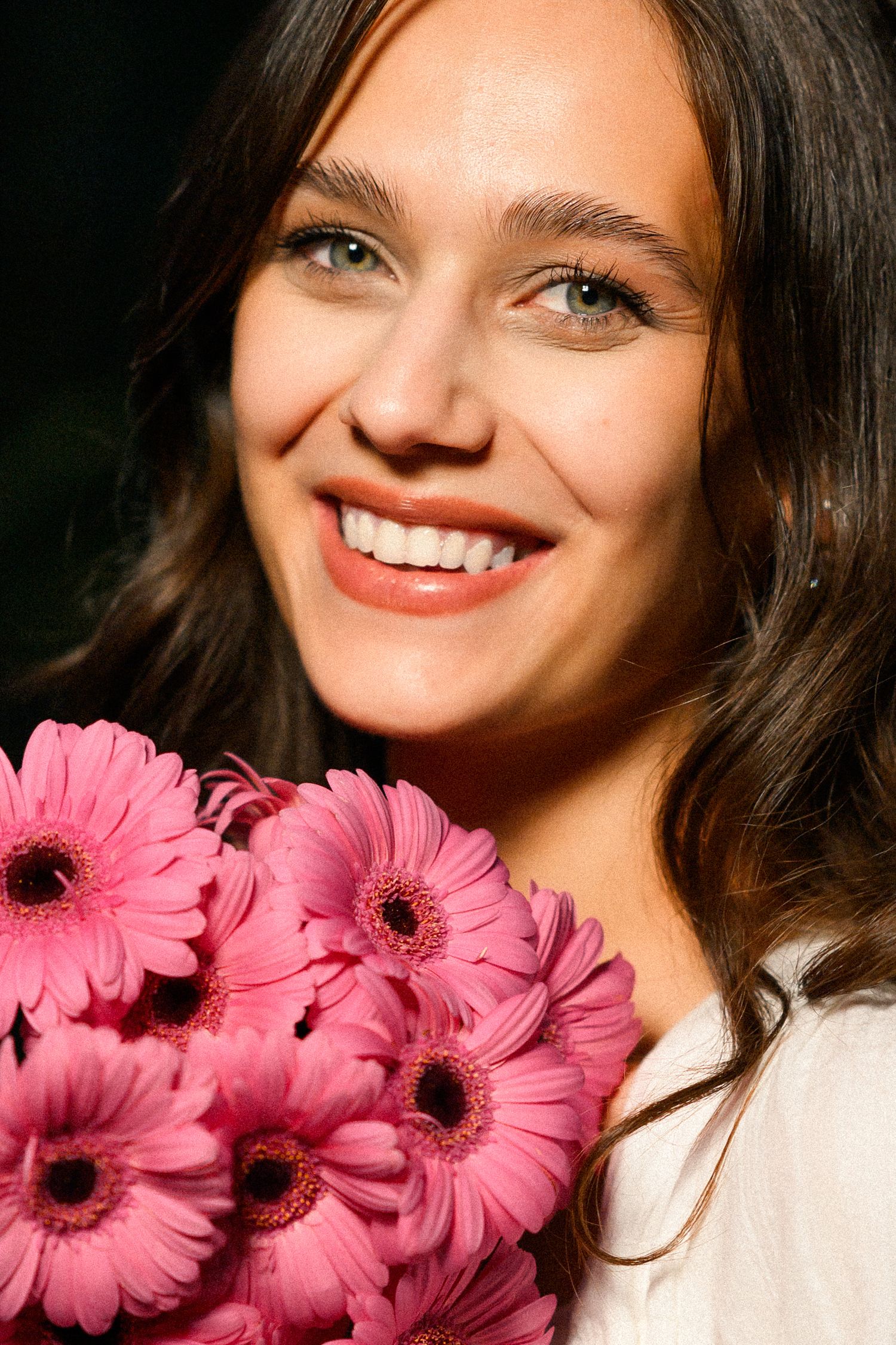 Smiling young woman with brown hair holding a bouquet of pink gerbera daisies against a dark background.