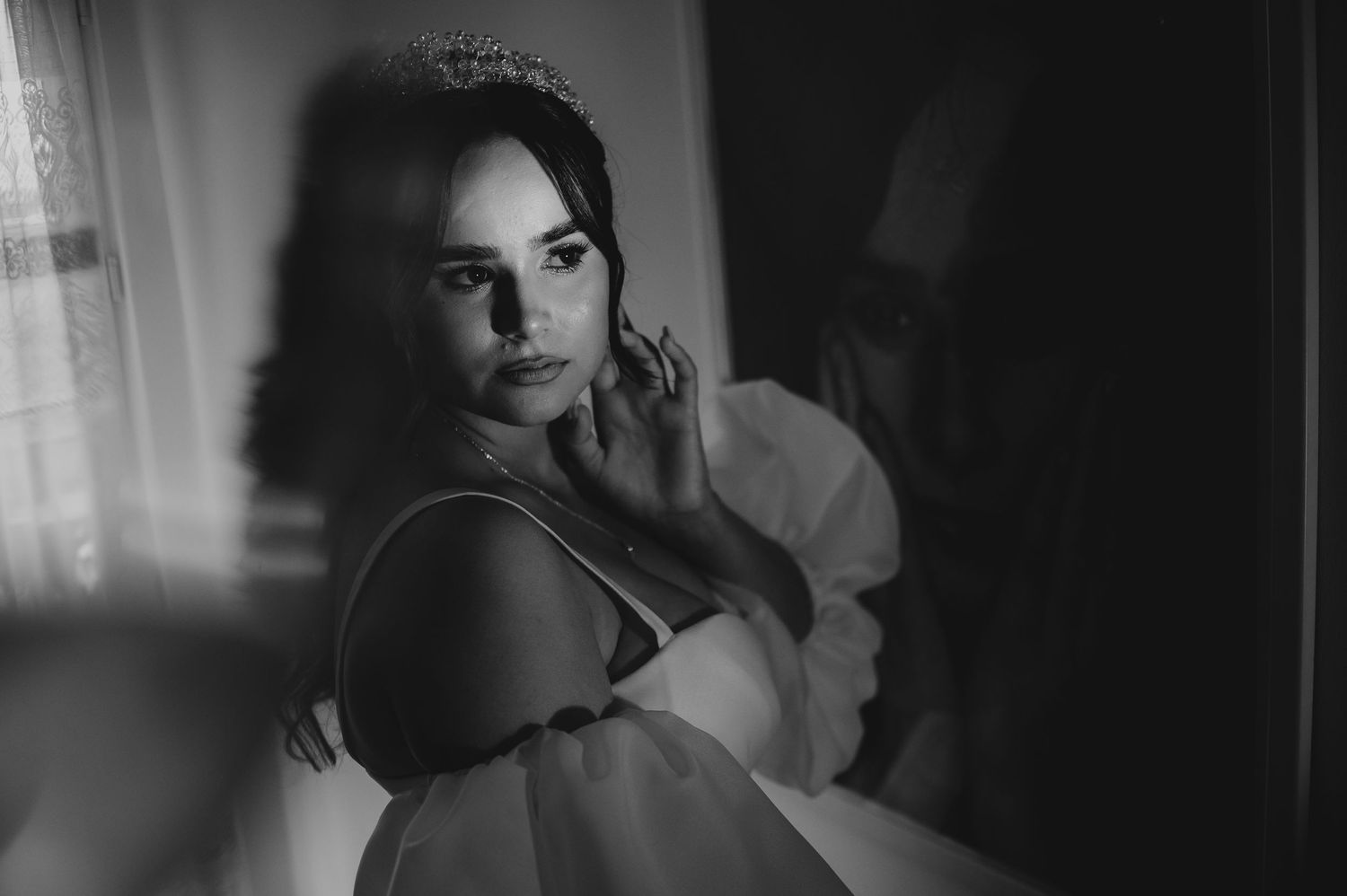 Black and white bridal portrait of a woman in a wedding dress and tiara posing thoughtfully by a window.