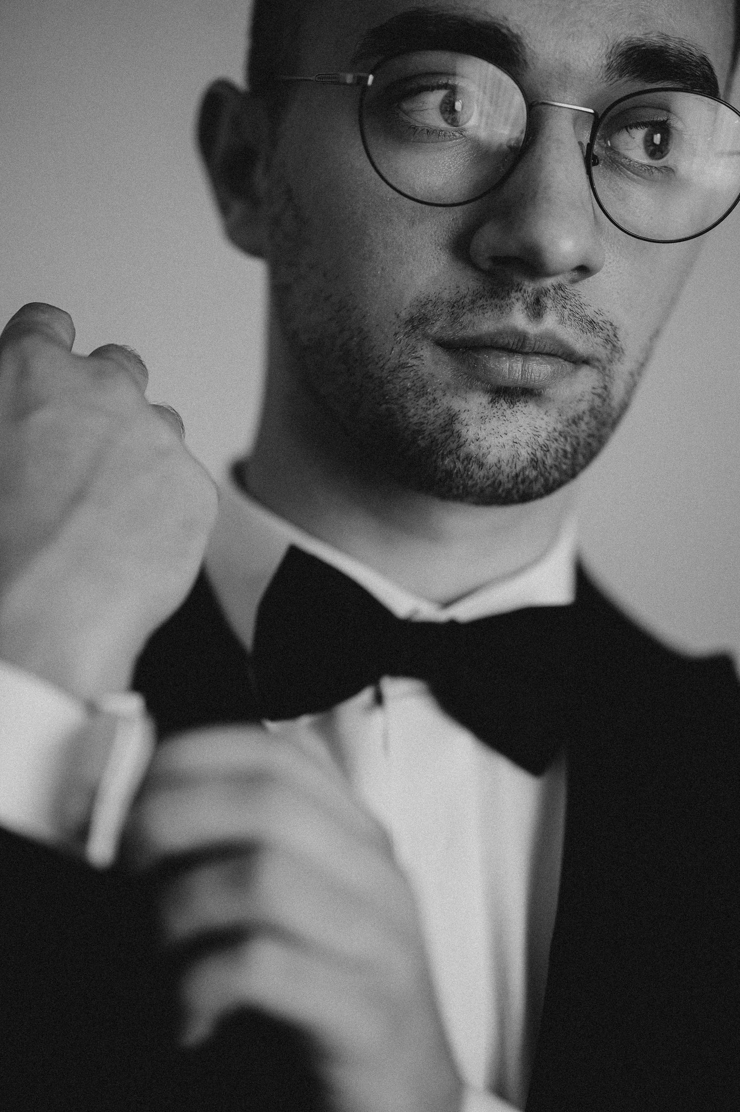 Groom in black tuxedo and bow tie adjusting cufflinks, wearing round glasses, black and white photo.