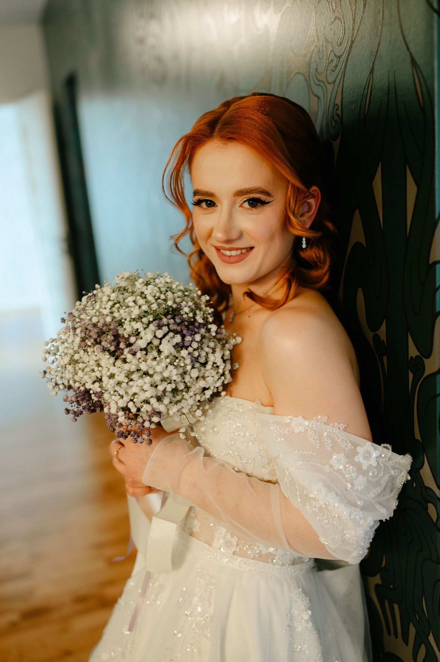 Smiling red-haired bride in off-shoulder white gown holding a baby's breath bouquet, leaning against a teal wall.