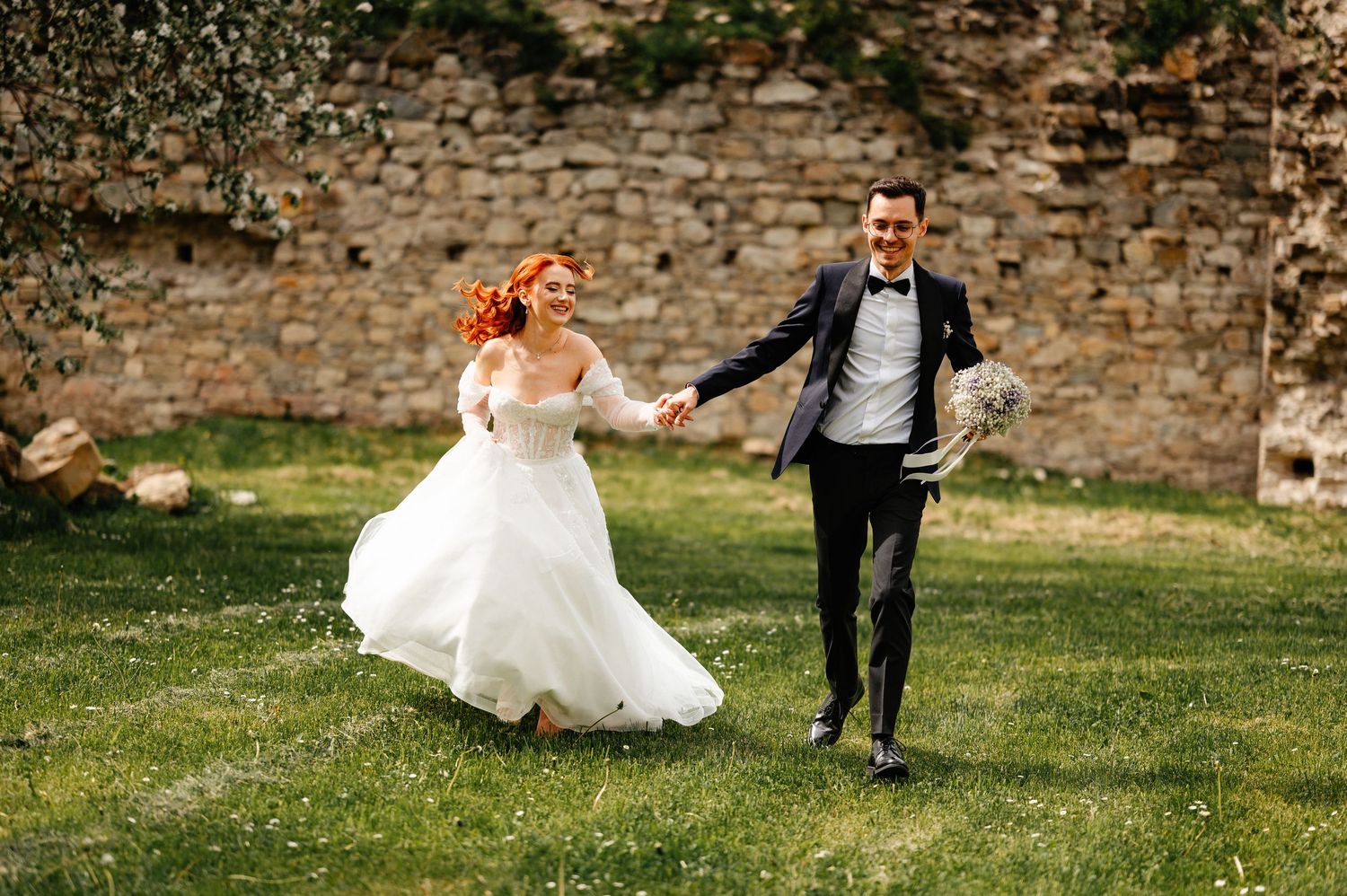 A joyful bride in a white gown and groom in a black tuxedo run hand-in-hand on green grass near a stone wall.