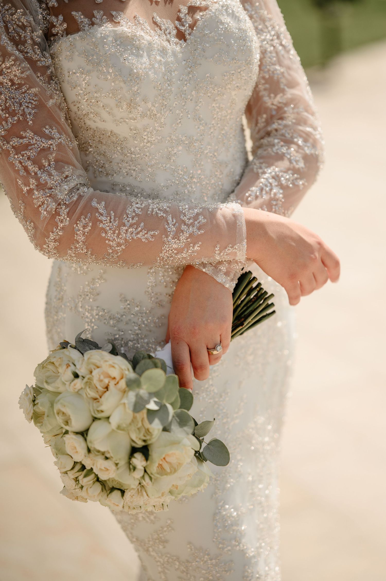 Close-up of bride in beaded long-sleeve lace gown holding elegant white rose and eucalyptus bouquet.
