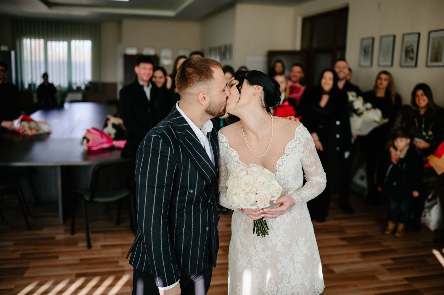 Bride and groom share a kiss at their wedding ceremony indoors, surrounded by guests, bride holding white bouquet.