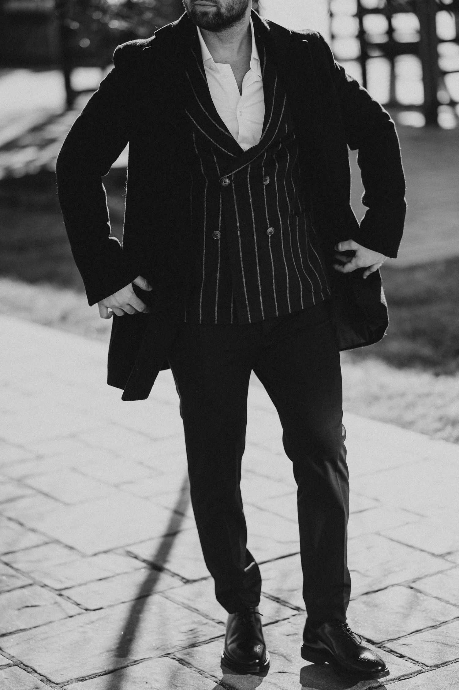 Black and white photo of a stylish man in striped vest and dark blazer posing outdoors on cobblestone.