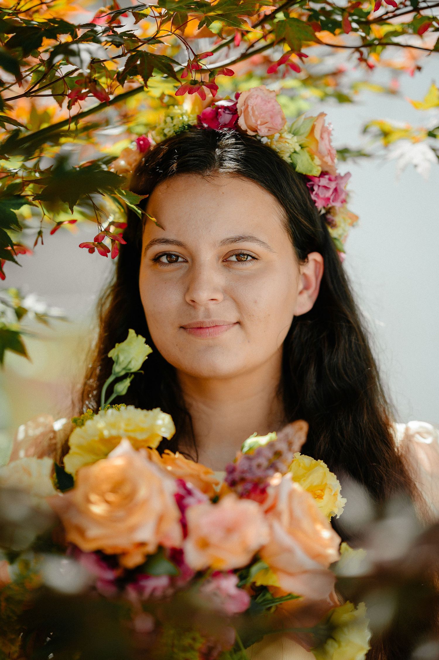Young woman wearing a floral crown with colorful roses and autumn leaves, holding a bouquet of peach and yellow flowers.
