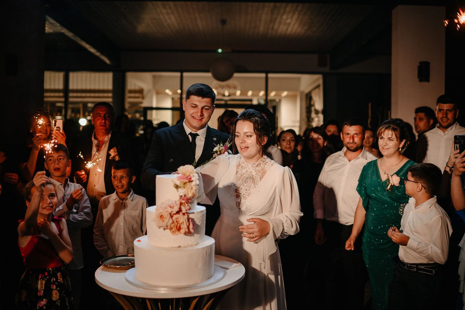 Bride and groom cutting wedding cake surrounded by guests holding sparklers at elegant evening reception.