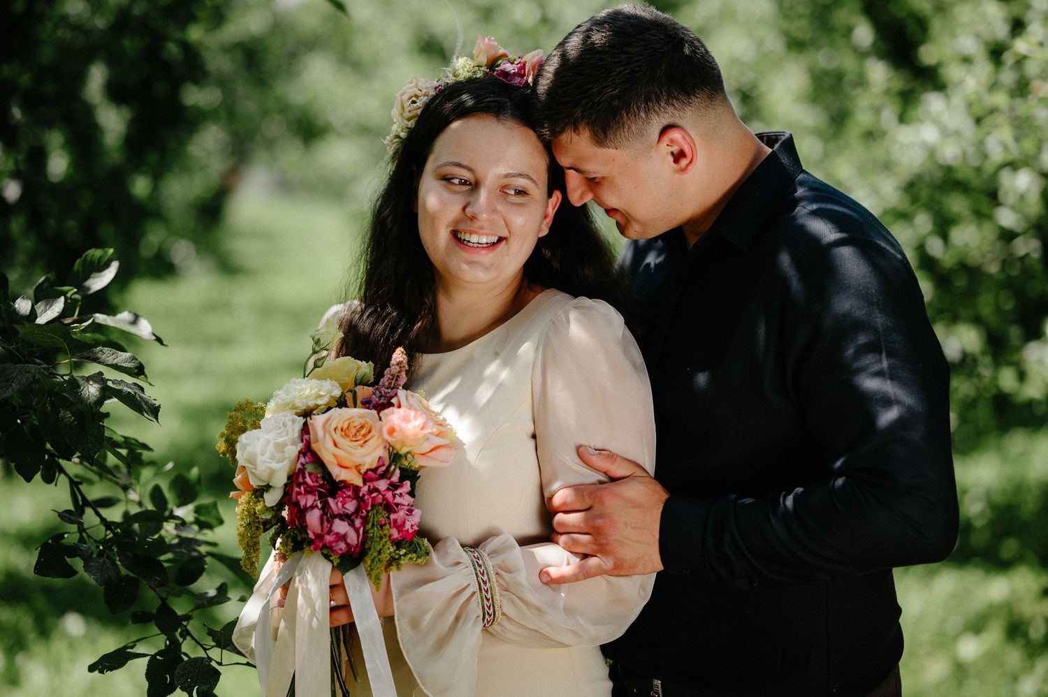 Smiling bride in white dress holding colorful bouquet embraced by groom in black jacket outdoors in green garden.