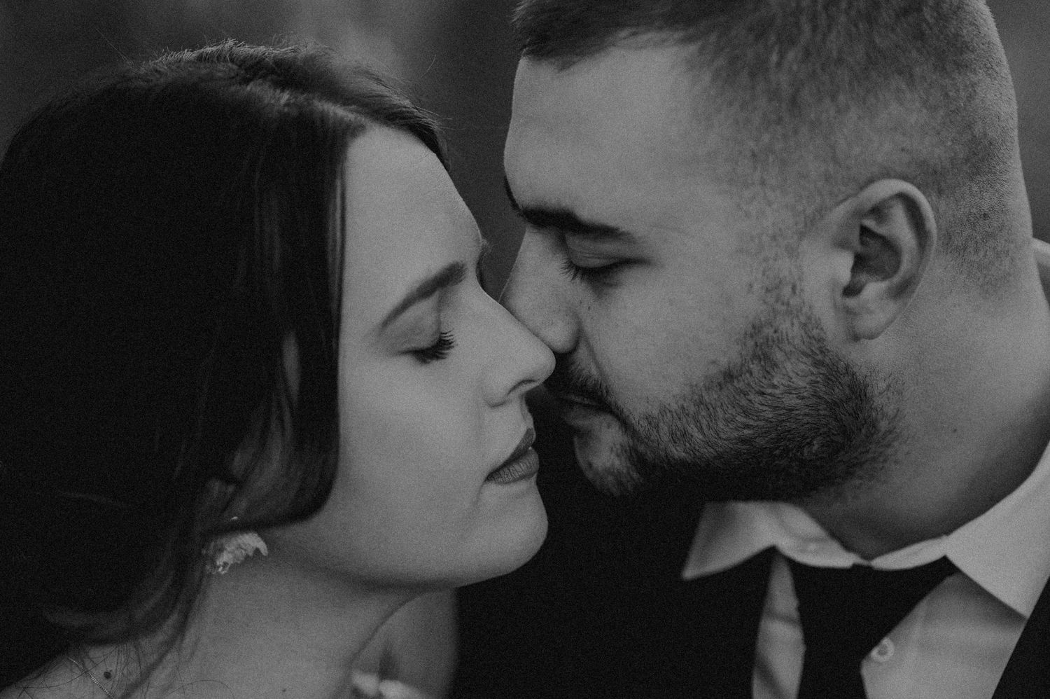Black and white photo of a couple sharing a romantic kiss, groom in suit, bride with dark hair.