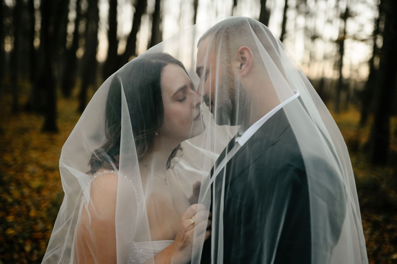 Bride and groom share a romantic moment under a wedding veil in an autumn forest at sunset.