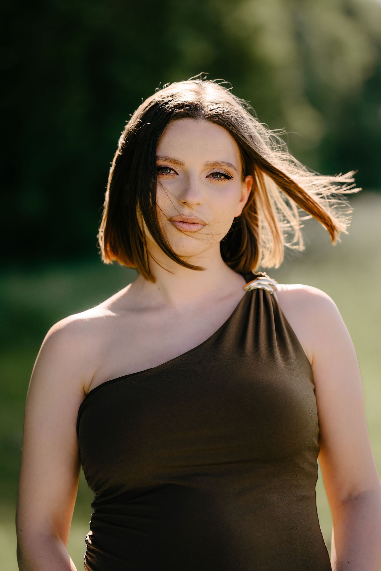Young brunette woman in dark one-shoulder dress with wind-blown hair outdoors.
