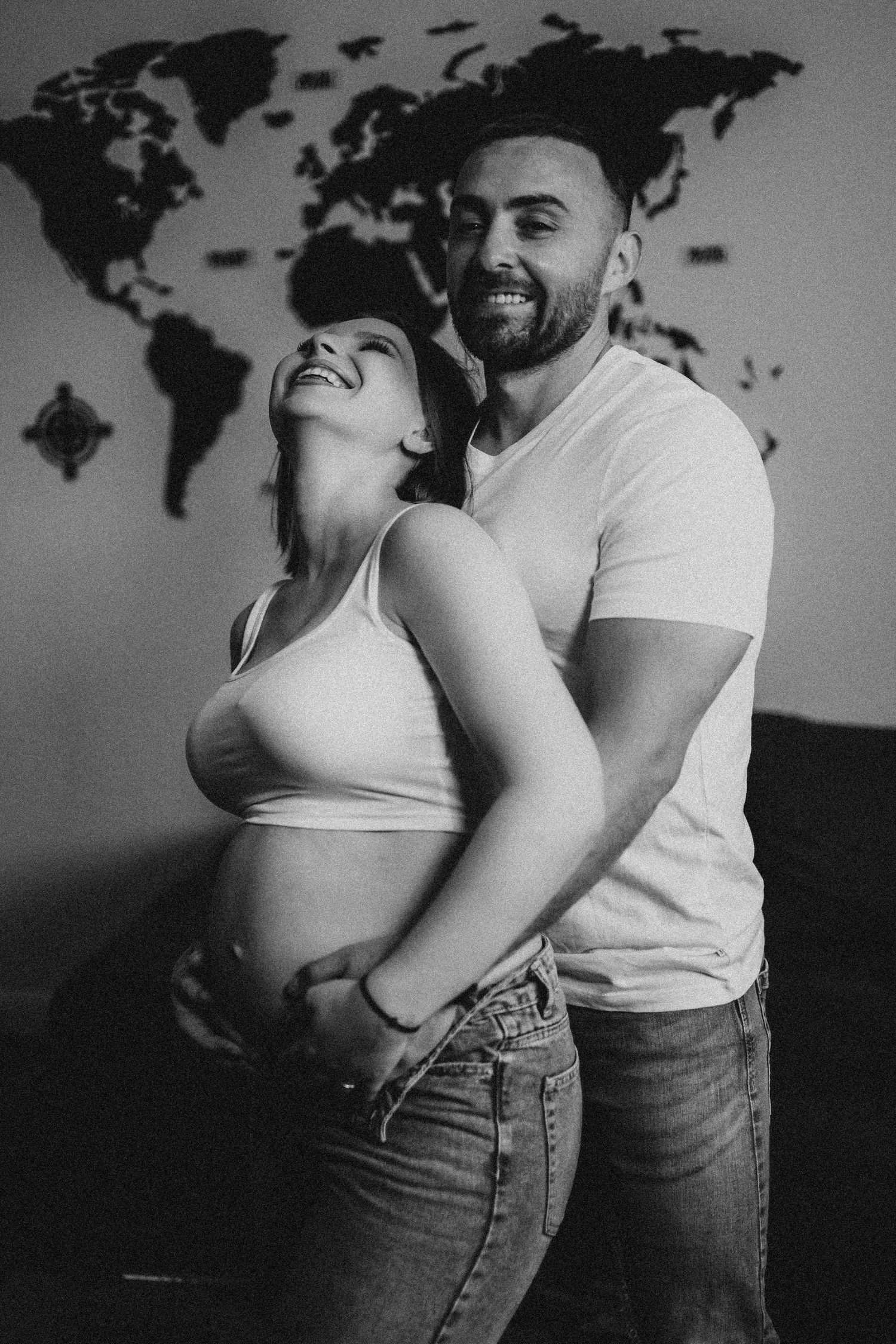 Happy couple embracing pregnant belly in black and white maternity photo with world map backdrop.