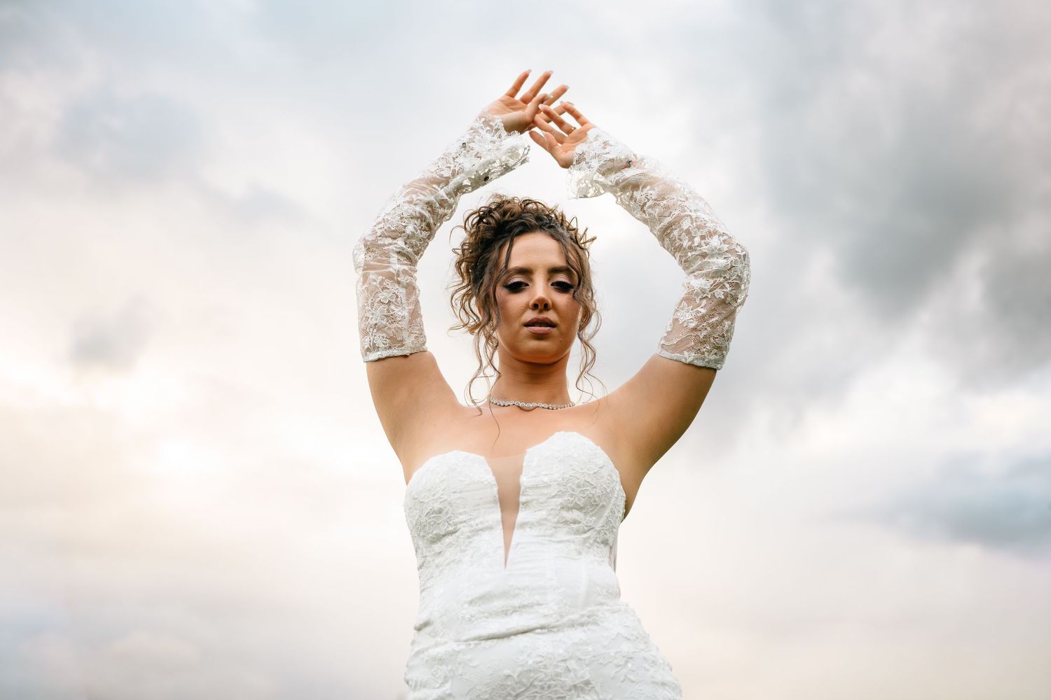 Bride in white lace gown raises arms gracefully against dramatic cloudy sky backdrop.