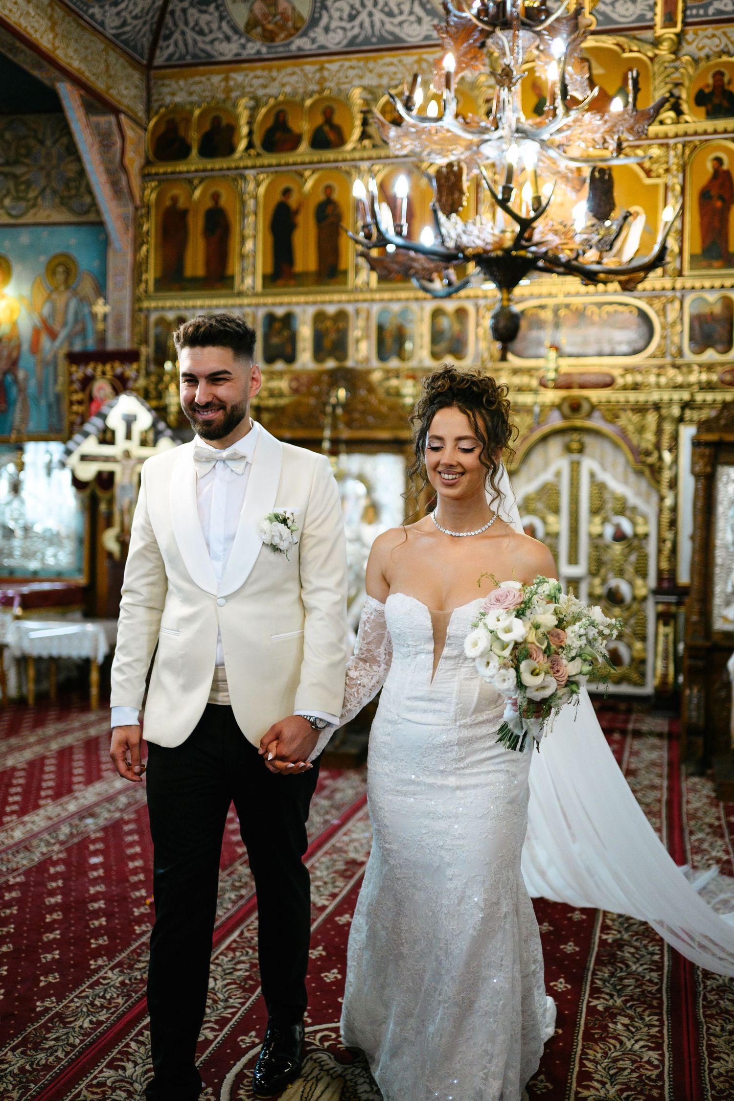 Bride and groom holding hands in ornate Orthodox church, bride in white gown with bouquet, groom in cream tuxedo.