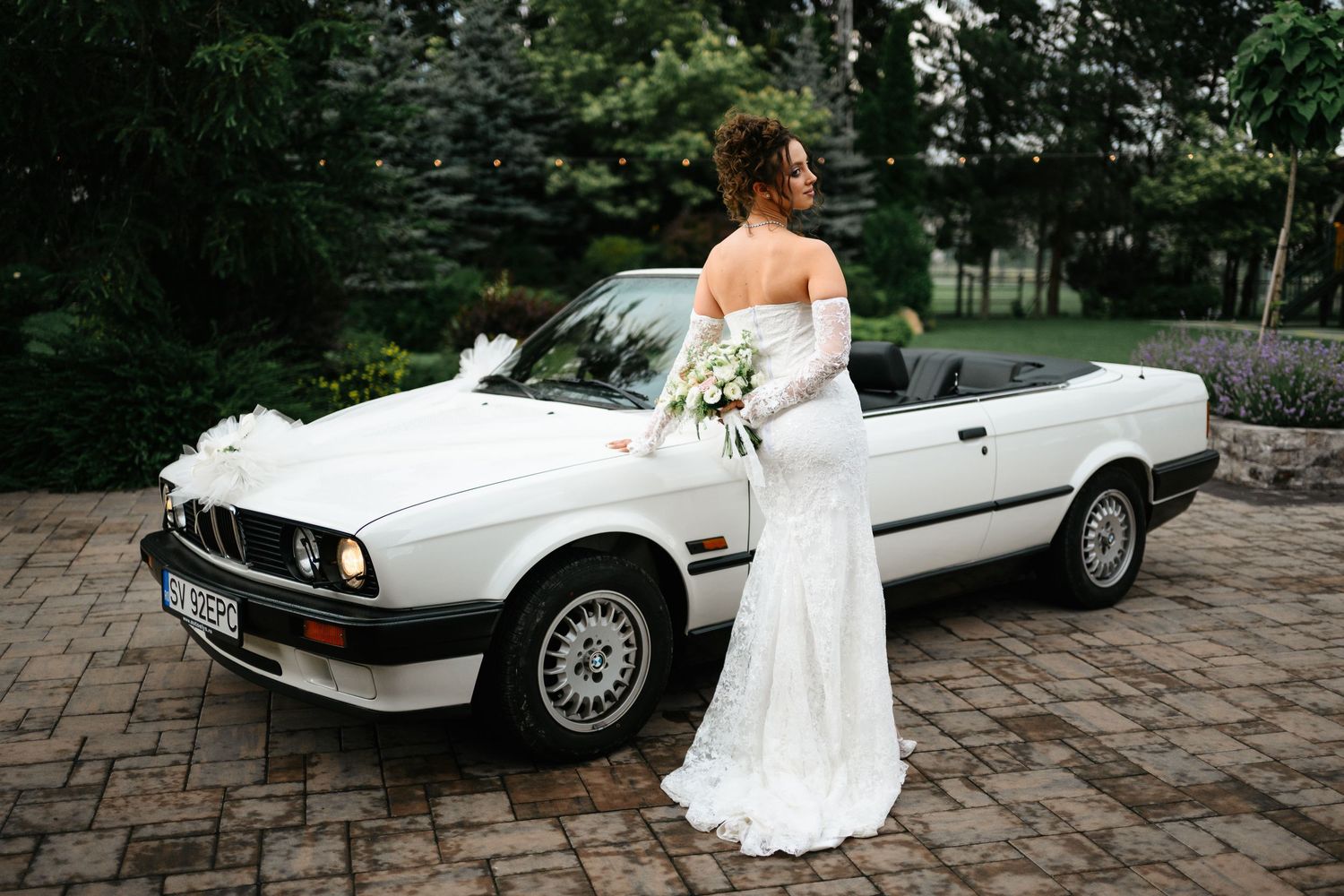 Bride in white off-shoulder gown holding bouquet beside a classic white BMW convertible decorated with ribbons.