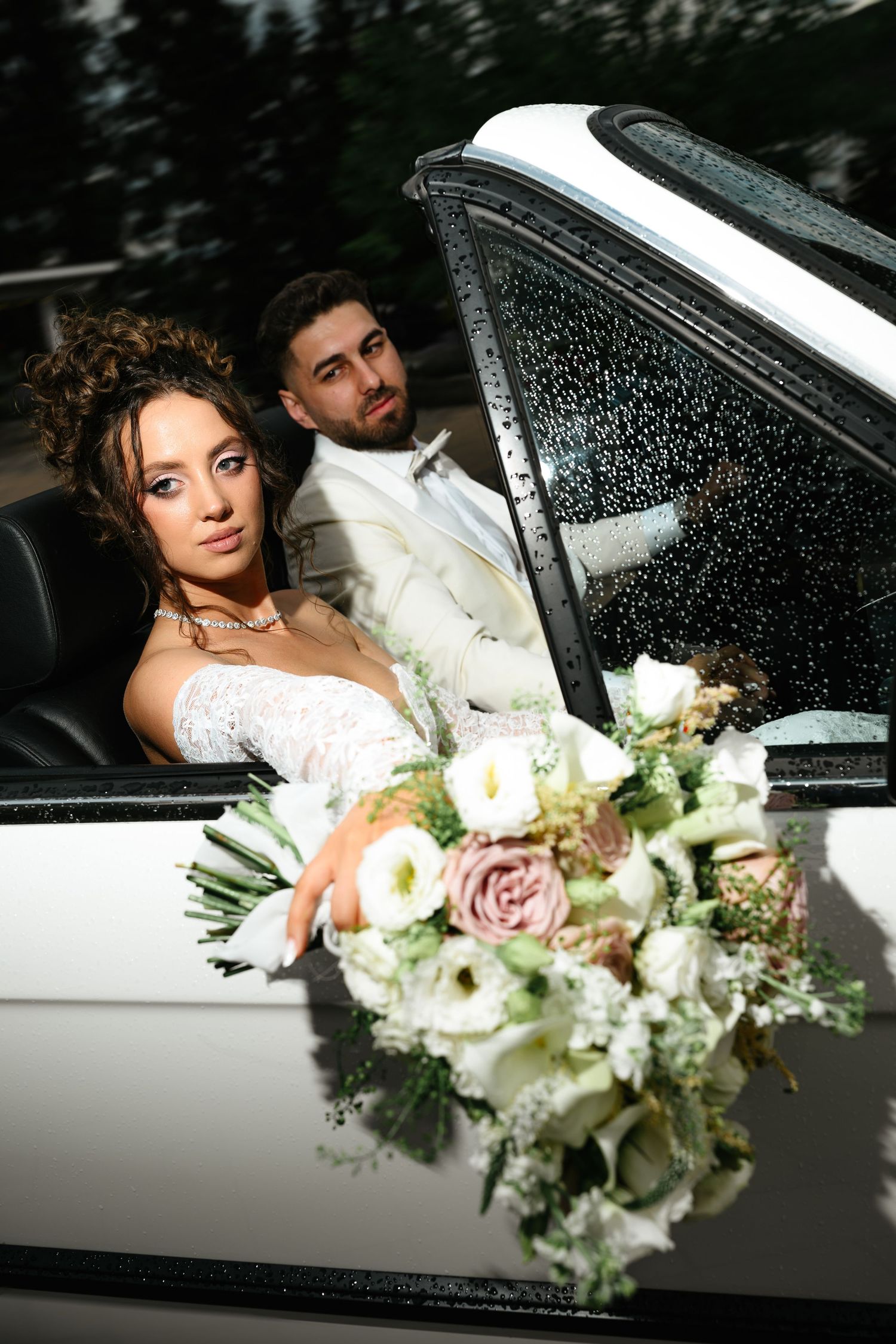 Bride and groom posing in a white convertible car, bride holding a bouquet of white and pink roses on their wedding day.
