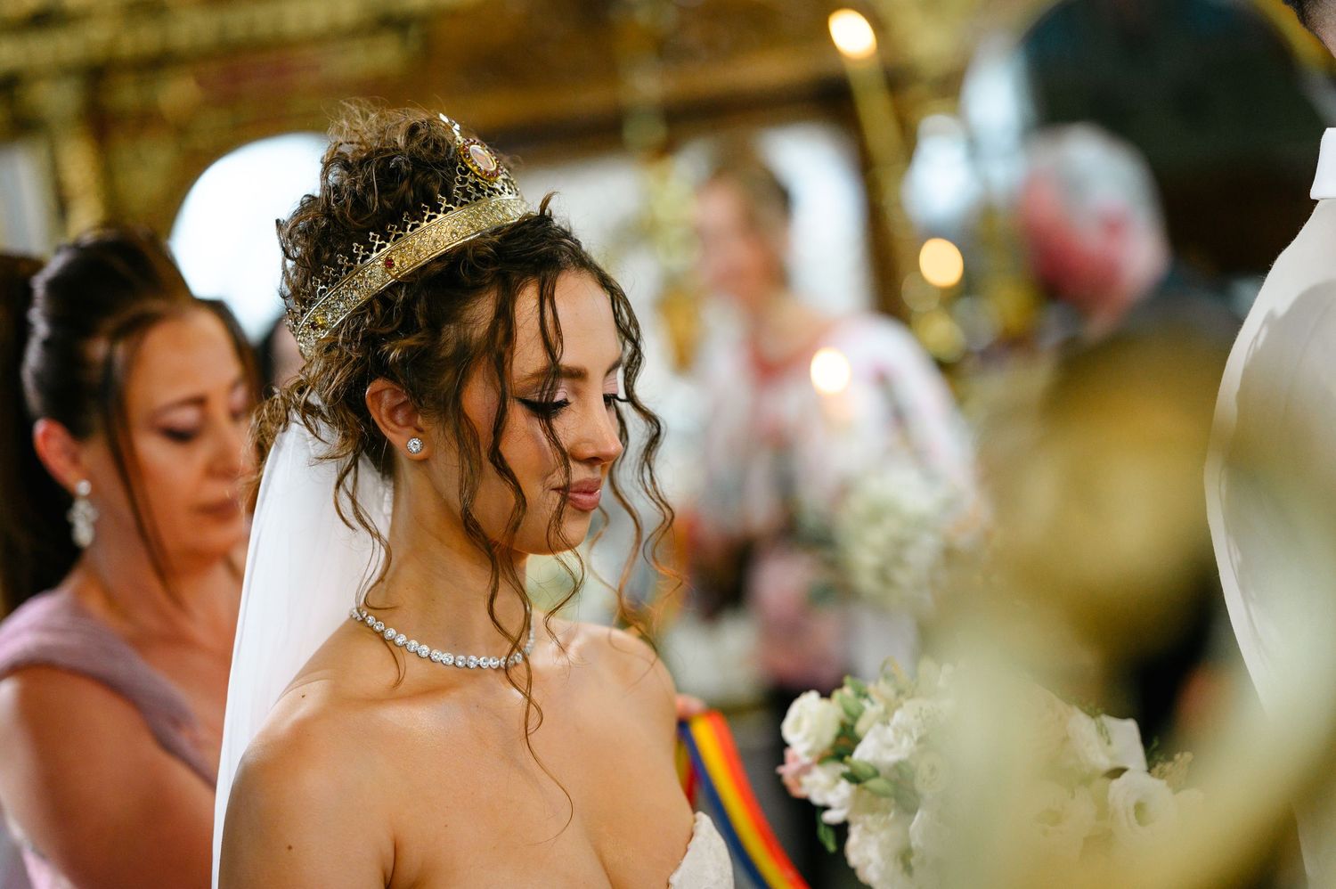 A bride wearing a tiara and veil looks down during a wedding ceremony surrounded by guests.