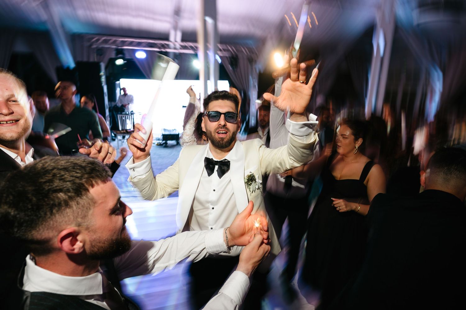 Groom in white tuxedo celebrating on dance floor with guests at wedding reception party.